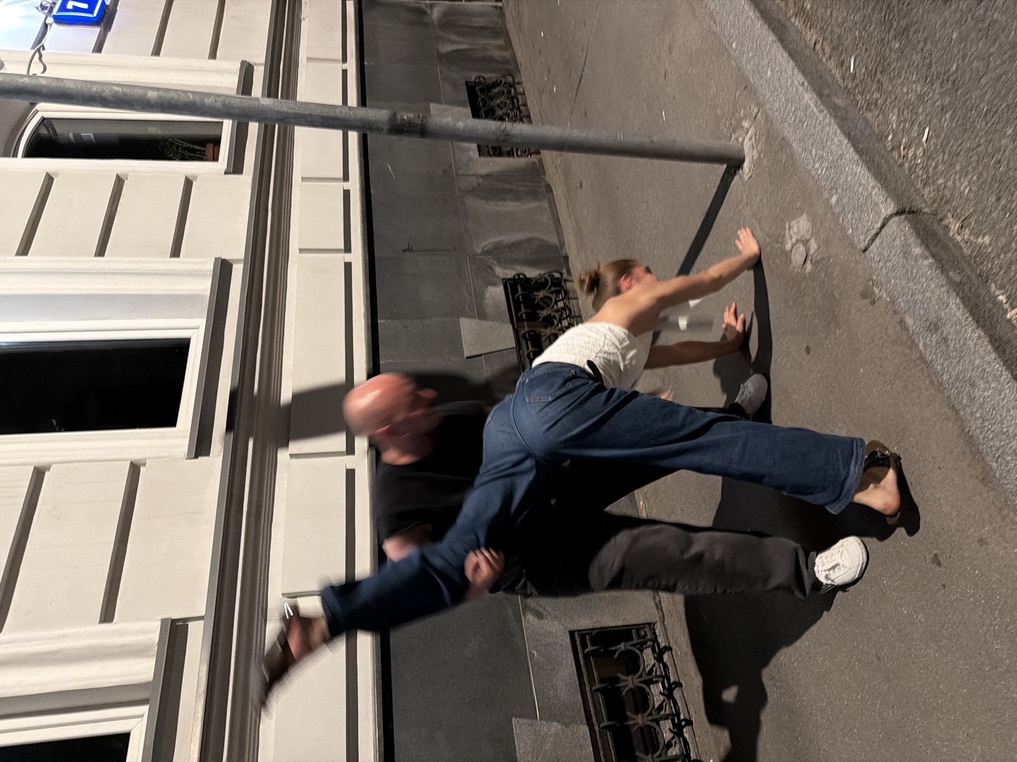 An unidentifiable man in a black t-shirt and dark pants is lifting an unidentifiable woman in a white top and blue jeans in a street scene. The woman is mid-air, with her legs extended. The man is standing on a sidewalk in front of a building with white facade and dark windows. A metal pole is visible to the right of the subjects. The scene appears to be at night, with artificial lighting illuminating the area. The overall impression is of a dynamic or playful interaction captured in a public urban space, possibly in Moscow, Russia, judging by the architectural style.