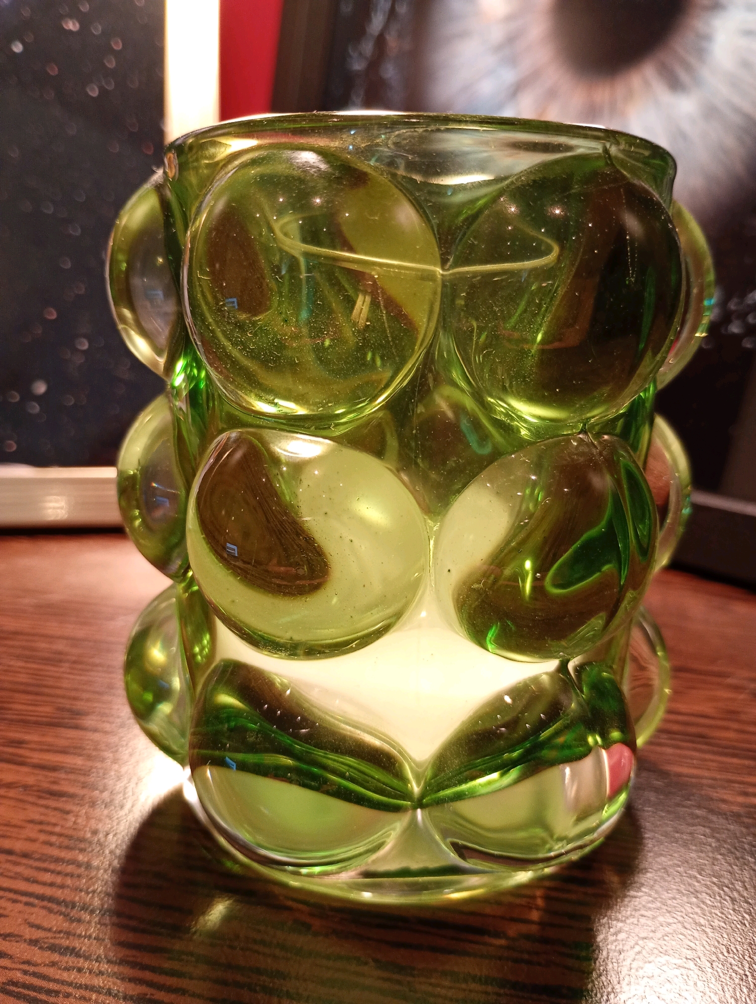 A close-up, low-angle shot features a green, textured glass candle holder resting on a brown wooden surface. The candle holder is cylindrical and made up of numerous bulbous, rounded protrusions that create a bubble-like effect. The glass is transparent and reflects light, creating highlights and streaks of darker green within its surface. The interior of the candle holder appears to be illuminated by a soft, white light. The background is out of focus, showing a dark area with scattered bokeh lights, a vertical stripe of red and yellow, and a blurred image of an eye. The scene does not contain any people, nor does it provide any information about its location in Bad Sachsa, Germany.