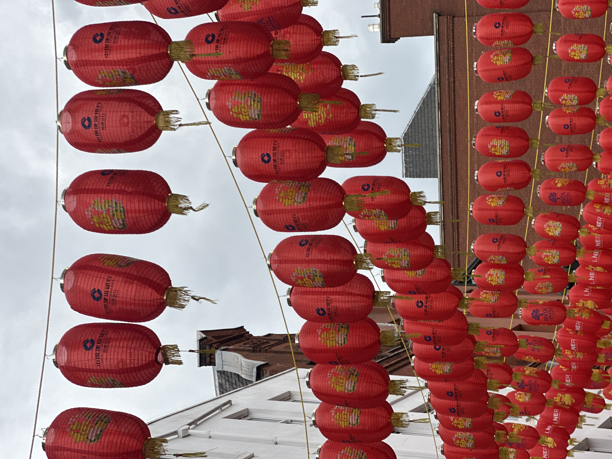 The image captures a street in London adorned with rows of red Chinese lanterns. The lanterns bear Chinese characters and the name of a bank, indicating a connection to Chinese culture. The buildings in the background have a mix of architectural styles. The sky is overcast.