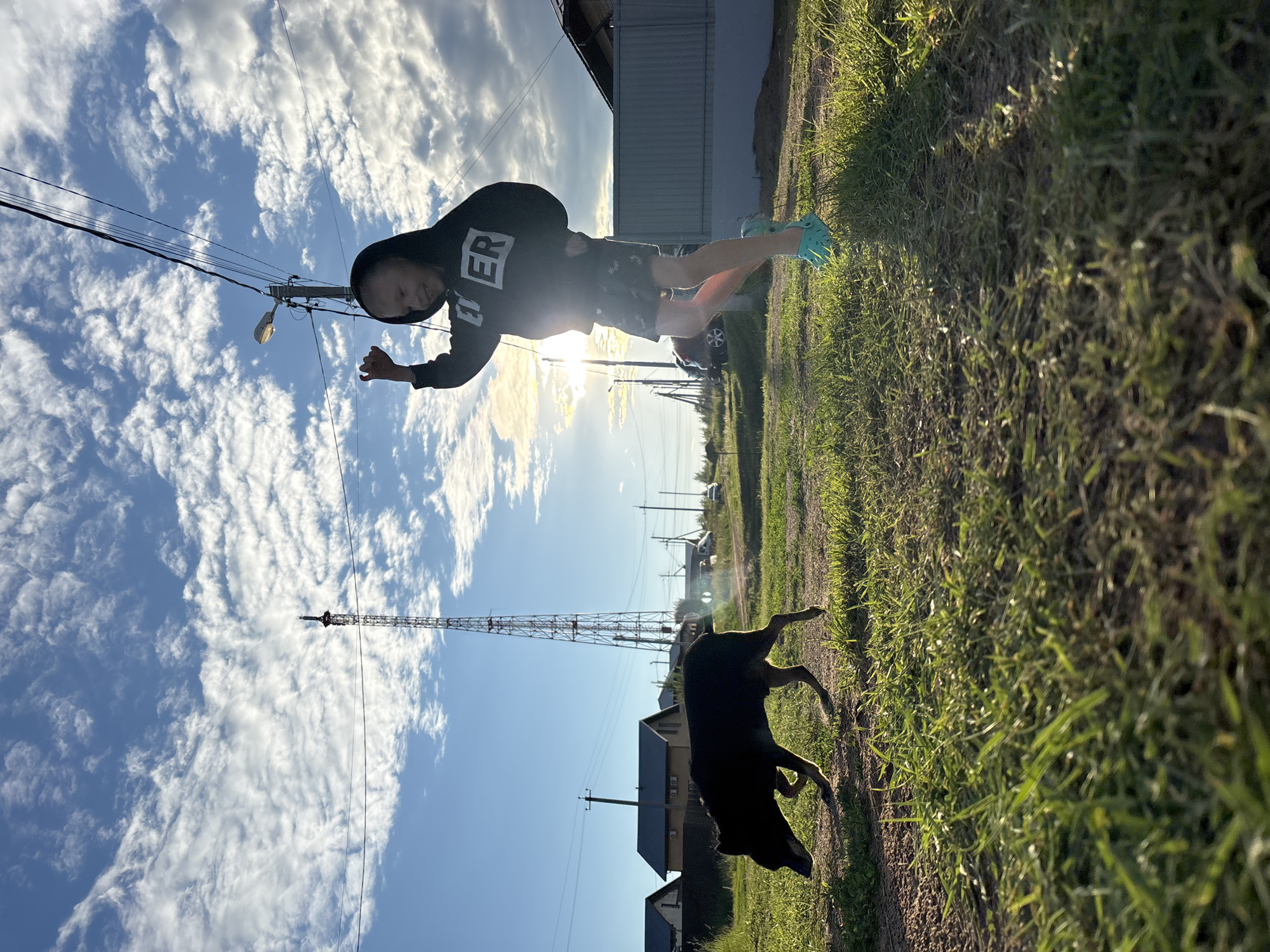 A young child, silhouetted against the setting sun, jumps in a grassy field with a black dog walking in the foreground. The child is wearing a dark hoodie with "ER" printed on it and patterned shorts. They are also wearing light blue clogs. A tall, lattice tower, likely a radio or cell tower, stands in the background. The sky is blue with scattered white clouds. Power lines and a street lamp are visible. In the distance, several houses and a gray fence can be seen. The scene appears to be a rural or suburban area, possibly on the outskirts of Chaikovsky, Russia, given the presence of similar infrastructure and housing styles. The lighting suggests it is late afternoon or early evening.