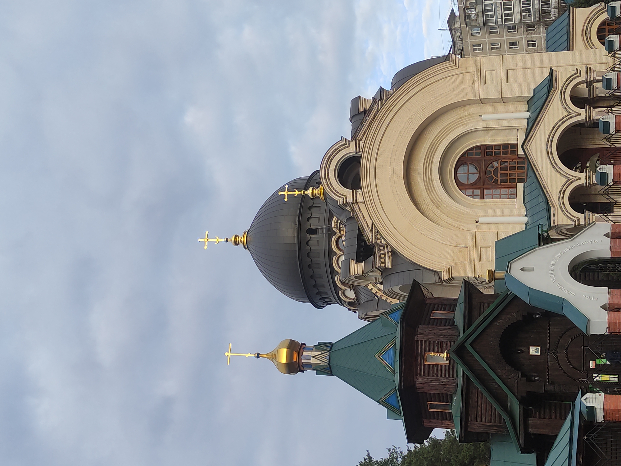 The image shows a complex of church buildings in an urban environment. The main architectural elements are:
1. A large, light-colored building with a prominent circular window and arched structures.
2. A dark-domed structure, possibly another part of the church, with golden crosses.
3. A wooden structure with a green roof and a golden dome.
A multi-story residential building is visible in the background. The sky is overcast. The location is in the city of Unknown, Unknown.