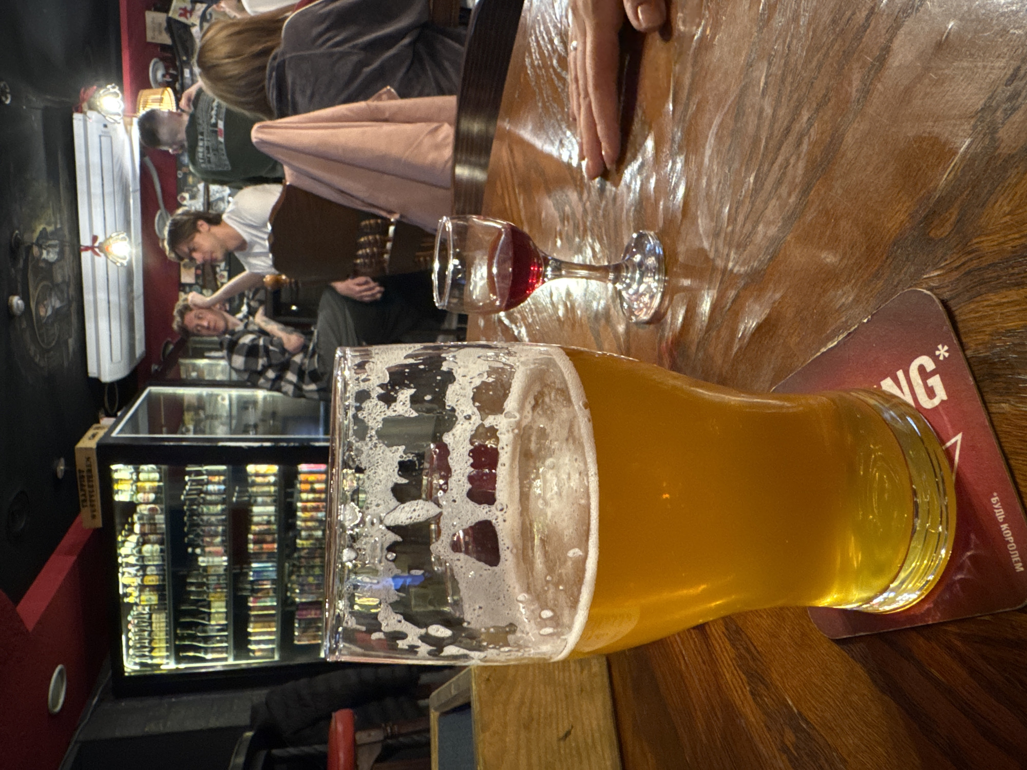 The image shows a table in what appears to be a pub or bar. In the foreground, a partially filled beer glass sits on a coaster. Next to it is a small wine glass containing a red liquid. The table has a wooden surface with a glossy finish.
Several people are visible in the background, including one wearing a plaid shirt. Behind them, there's a glass-fronted display case filled with various items, possibly bottles or other products. On the left side, the wall is painted a dark color with a lighting fixture on the ceiling. The location is identified on the coaster as Chaikovsky, Russia.