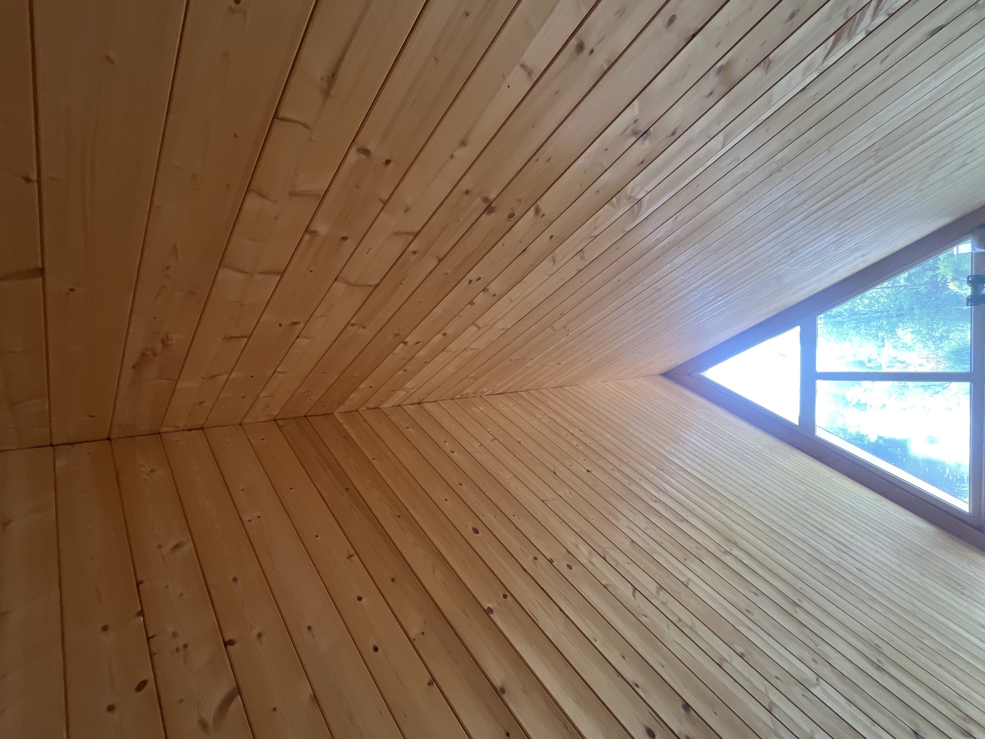 The image shows the interior of a wooden A-frame structure, likely a room or cabin, with a triangular window at the apex. The walls and ceiling are clad in light-colored wooden planks, running parallel to each other and meeting at the peak. The window is clear and allows light to enter, illuminating the interior and revealing a blurred view of green foliage outside, suggesting trees. The overall impression is one of a rustic and natural aesthetic. This scene is located in Vasilyevo, Russia.