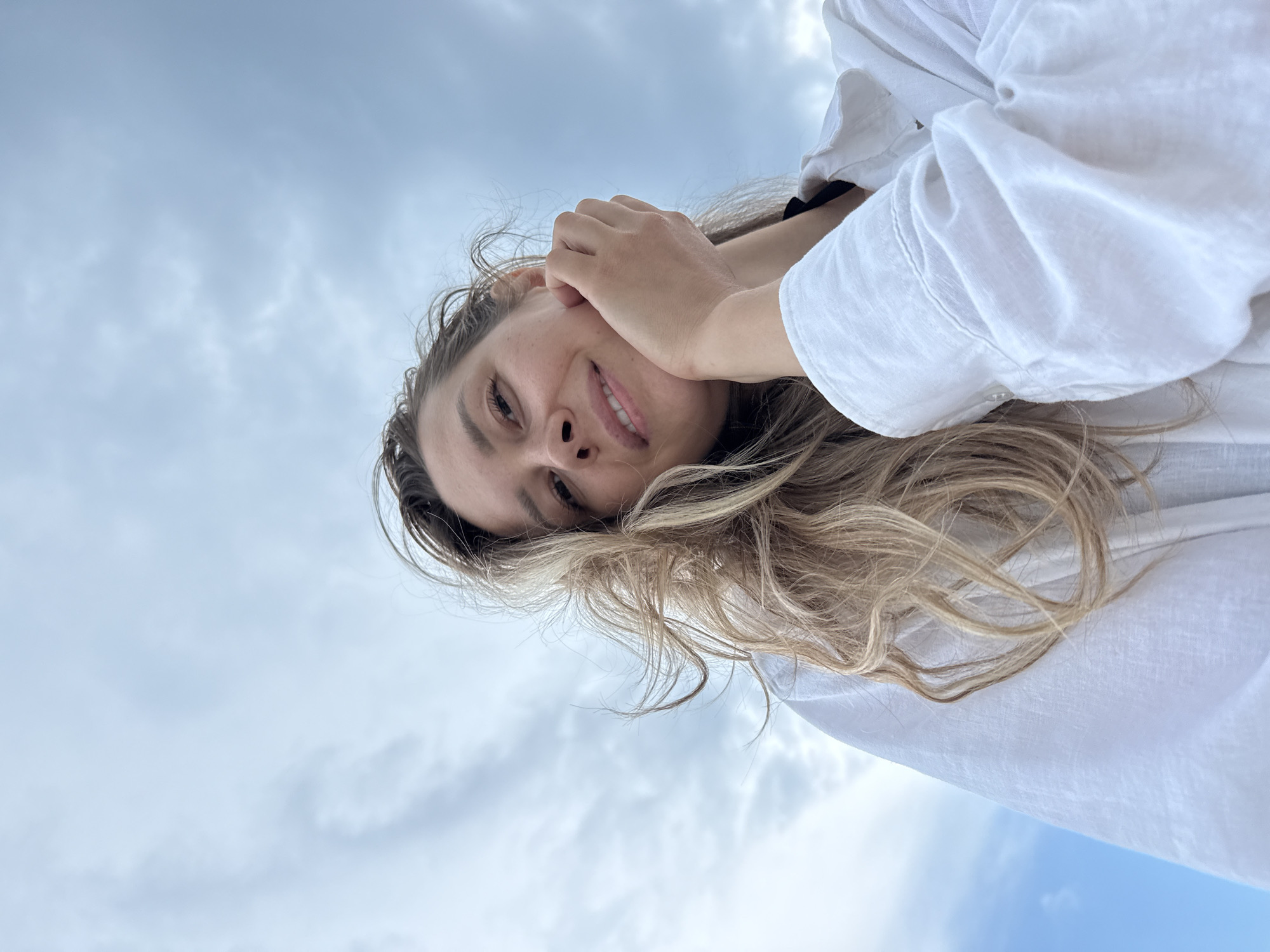 A low-angle, medium shot features a young woman outdoors against a cloudy sky. She has blonde, wavy hair that is partially obscuring her face. The woman is wearing a white collared shirt with the sleeves rolled up. Her right hand is resting on her chin, and she is smiling slightly at the camera. The background is dominated by a hazy sky with a few patches of lighter blue. The scene appears to be set in the city of Nebug, Russia.