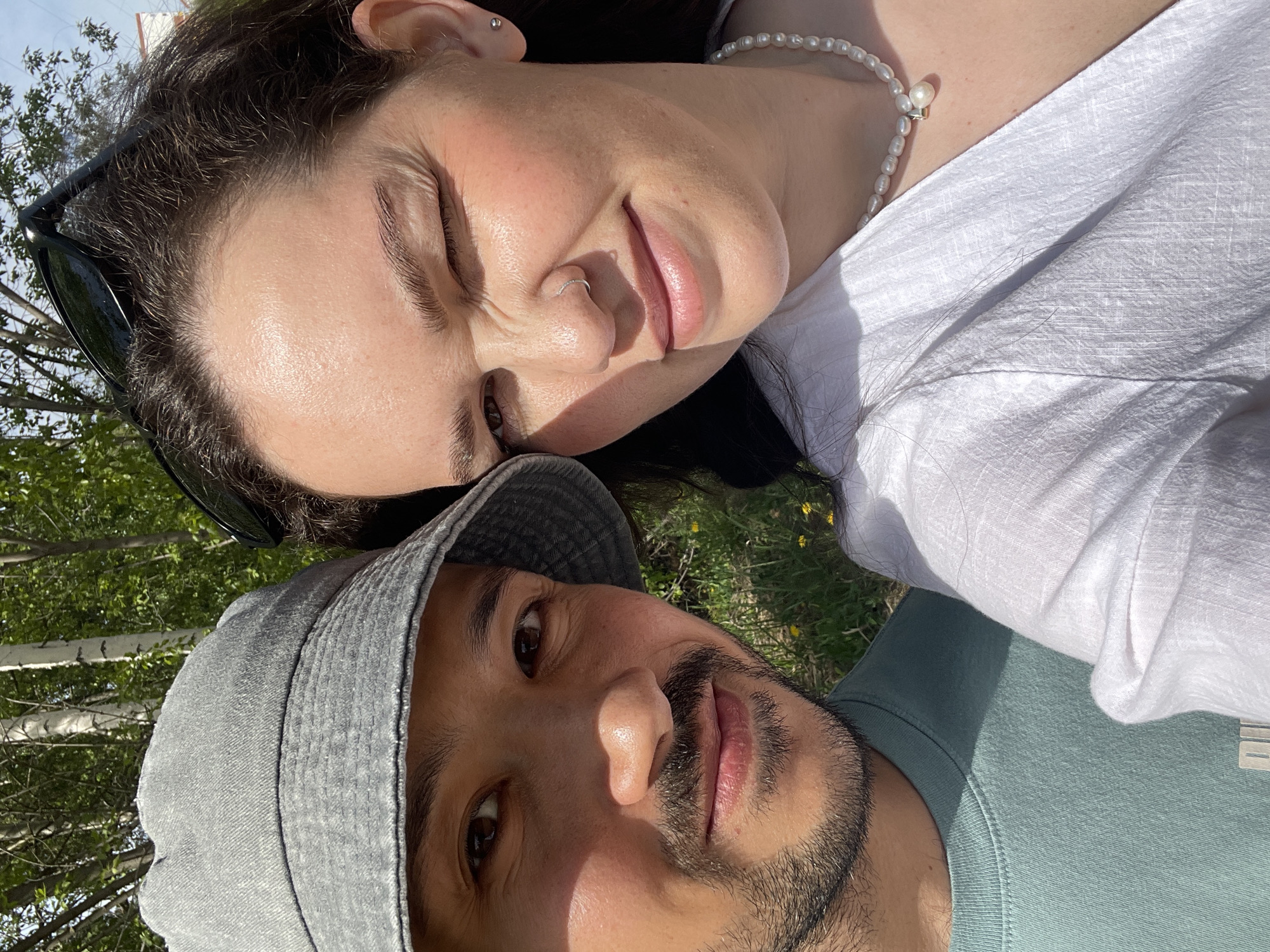 Two people are closely posed together, with one person wearing a grey bucket hat and a light green t-shirt and the other wearing a white top and a pearl necklace. They appear to be outside, possibly in a park, given the visible trees and grass in the background. Sunglasses are resting on the top of the head of the person wearing the white top. The image lacks specific location information to verify its location in Izhevsk, Russia.