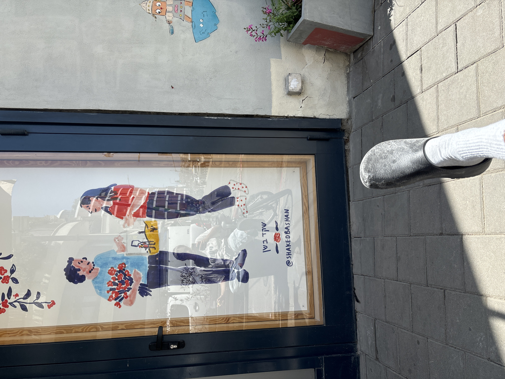 The image captures a street scene, seemingly in Tel Aviv, Israel, as indicated by the Hebrew text visible on a storefront window. A pair of dark blue doors are evident, with a large window in between. The window features a poster illustration showing two figures holding flowers, with the name "SHAKED BASHAN" and an "@shakedbashan" handle next to the text.
The outside wall is light gray, decorated with drawings, and has a light gray pot on the upper right corner. A light gray street is on the right of the image.
A person's foot, wearing a white sock and a gray shoe, is positioned in the upper-right corner of the image.