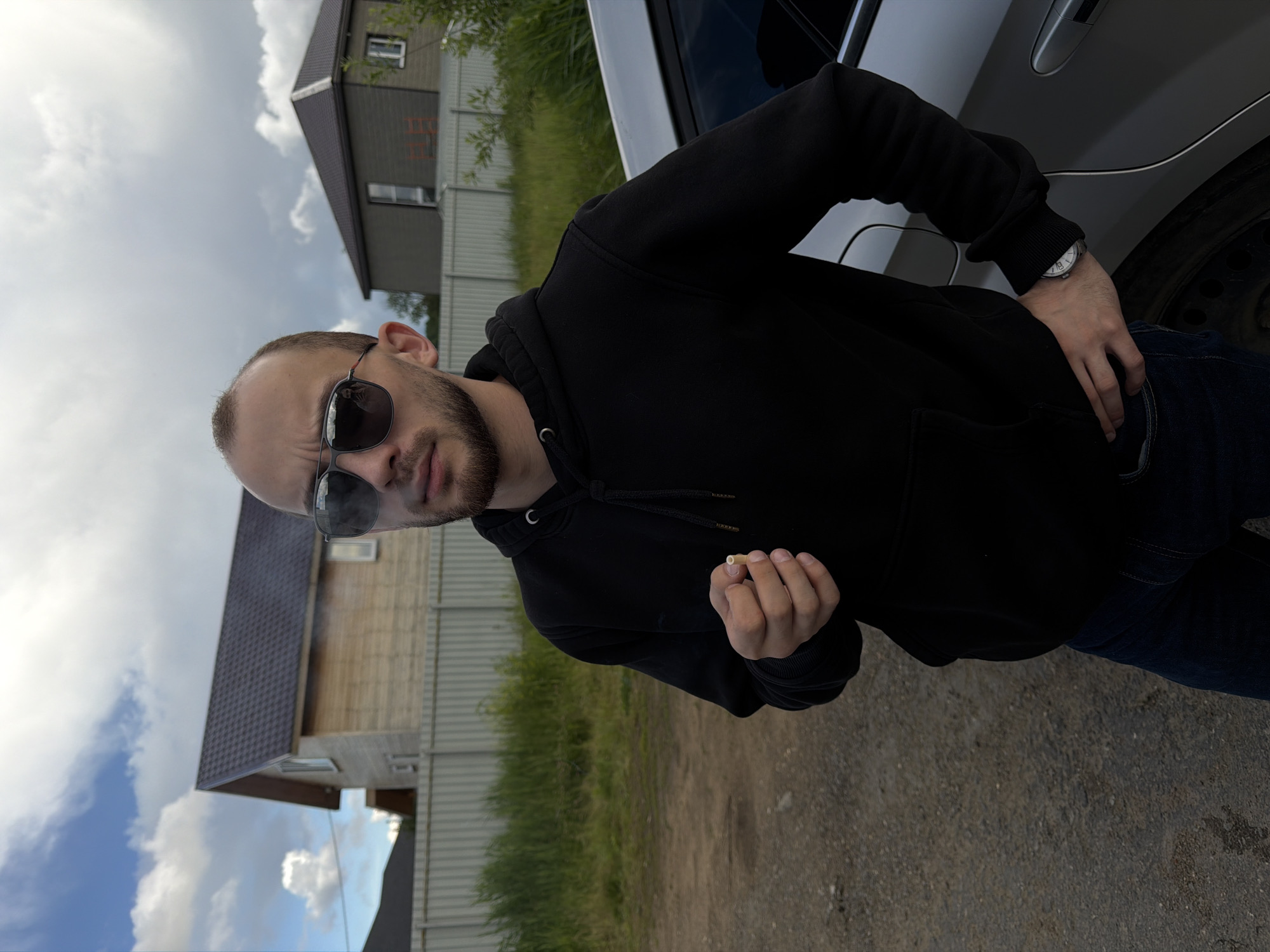 This is a vertically oriented, medium shot of a man standing next to a car. The man, with a beard and wearing sunglasses and a black hoodie, is holding a cigarette in his right hand. He appears to be in an outdoor setting with a building and a grassy area in the background. The sky is cloudy, suggesting it might be an overcast day. The man's expression is difficult to discern due to the sunglasses, but his posture suggests a relaxed or perhaps thoughtful demeanor. The car appears to be a silver sedan. The overall lighting indicates it is daytime.
