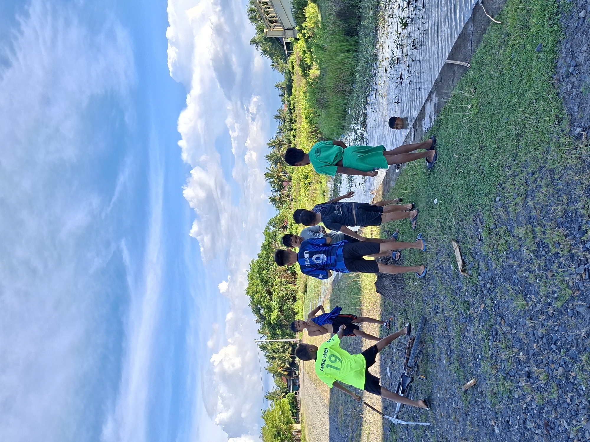 In Keudegeudong, Indonesia, a group of six boys are gathered near a waterway. One boy is in the water, with only his head visible. The others stand on a grassy bank. One boy wears a bright green shirt with the number 19 and the name "LAMINE YAMAL" printed on the back and is on a bicycle. Another boy wears a blue jersey with the name "THURAM" and the number 9 on the back. The environment includes a bridge in the background, a body of water, and a clear sky with clouds.