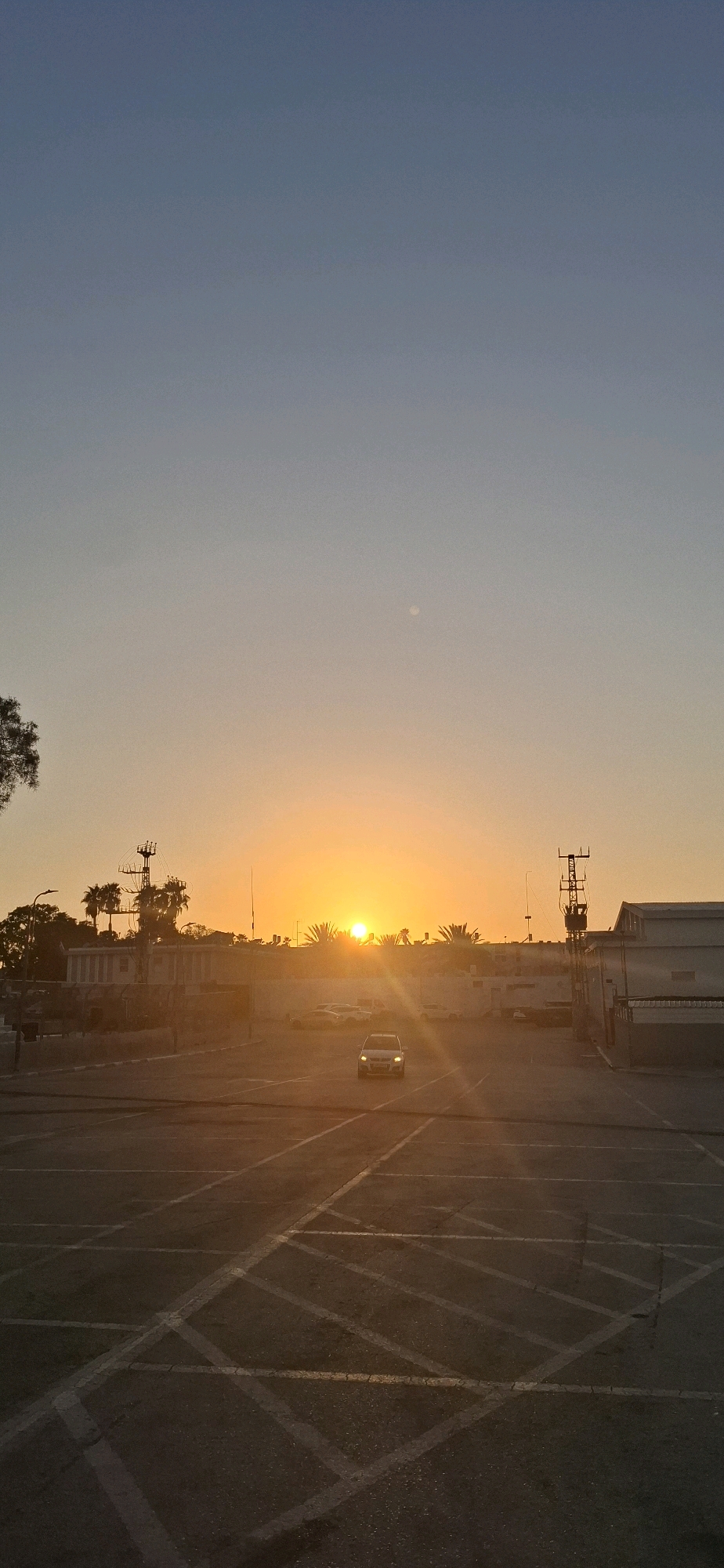 The image depicts a sunset scene in Yerocham, Israel, possibly an industrial area. In the foreground, a dimly lit asphalt parking lot is visible with painted parking lines. A white car is driving towards the viewer in the middle of the parking lot, its headlights on. In the background, a line of buildings and palm trees are silhouetted against the bright orange and yellow hues of the setting sun. The sky transitions from a clear, pale blue at the top to warm tones near the horizon. A faint circular shape, possibly the moon or a planet, is visible in the upper portion of the sky. Two utility poles with electrical equipment are also present, one on the left and another on the right side of the frame. The overall atmosphere is tranquil, with the warm light of the sunset casting long shadows.