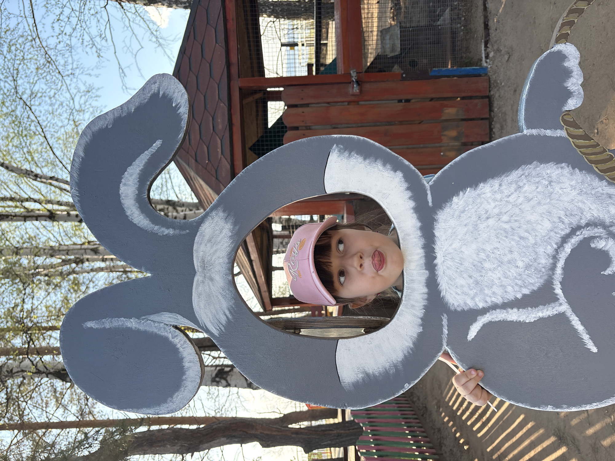 A medium shot depicts a young girl in Chaikovsky, Russia, sticking her tongue out through a cutout in a painted gray wooden rabbit. The girl wears a pink baseball cap. Behind her is a wooden structure resembling a playhouse or small animal enclosure with wire mesh visible. Trees with sparse leaves are visible in the background, along with a blue sky. A section of a striped bench is visible at the bottom of the frame. The rabbit cutout is textured with white paint suggesting fur, and it's positioned such that the girl's face fills the hole where the rabbit's head would be. The photo appears to be taken during daylight.