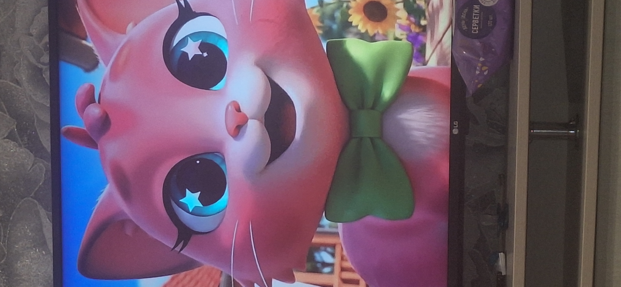 The image is a close-up shot of a pink animated cat with large blue eyes featuring a white star shape. The cat has a green bow tie around its neck and appears to be outdoors with a blurred background of sunflowers and greenery. The cat's expression is happy, with a wide smile and its mouth open. Below the cat, the top of a black television screen with an "LG" logo is visible, and on the shelf below the screen, there is a package of wet wipes. The image does not contain any identifiable landmarks or features that would allow for its location to be determined in Bila Tserkva, Ukraine, or any other city.