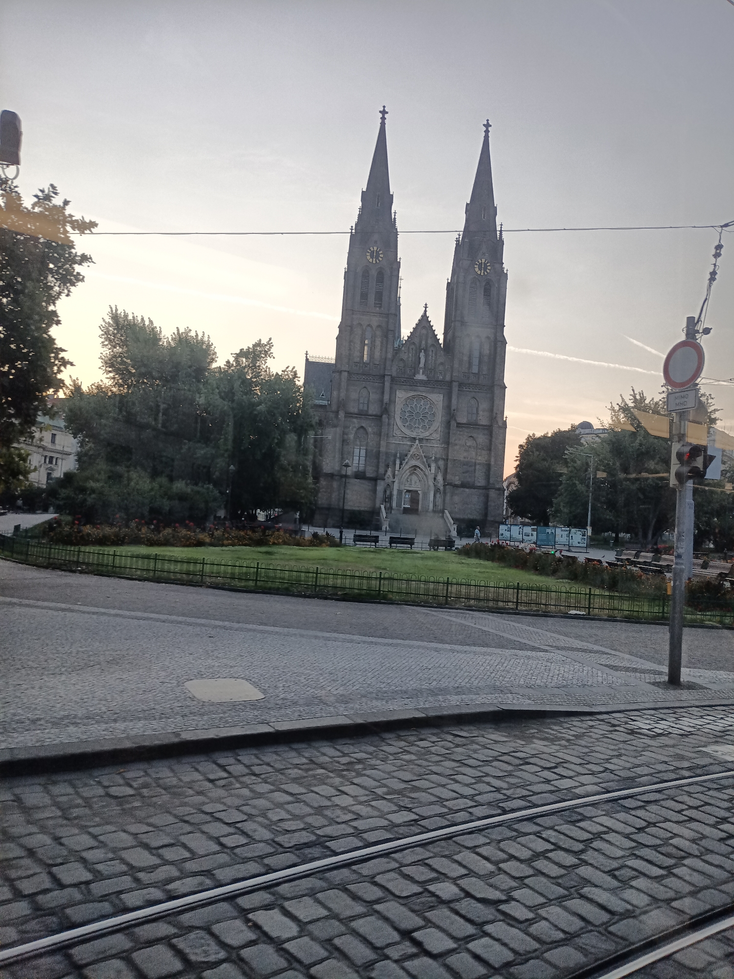 The image shows the Basilica of St. Peter and St. Paul in Vyšehrad, Prague, Czechia. The basilica is a Gothic Revival church with two prominent spires and a rose window above the entrance. It is situated on a hill overlooking the city. In the foreground, there is a cobblestone street with tram tracks. A fence surrounds a green lawn in front of the church, with flowerbeds and trees. A traffic light with a red signal is visible on the right side of the image, along with a circular traffic sign. The sky is overcast with some faint contrails. The overall scene depicts a peaceful urban landscape with historical architecture.