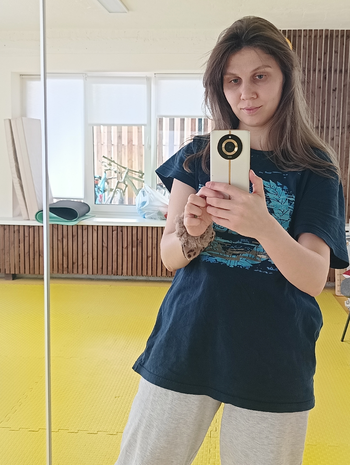 A young woman is depicted in a mirror selfie, holding a smartphone with a distinctive circular camera module in both hands. She has long, wavy brown hair, parted to the side, and looks directly into the camera with a neutral, calm expression. She is wearing a dark blue t-shirt with a light blue graphic design across the chest and light gray sweatpants. A scrunchie or hair tie is visible on her left wrist.
The indoor setting, reflected in the mirror, features a large window in the background with white roller blinds partially drawn. Outside the window, a wooden fence and a bicycle can be seen. On the windowsill, several items are placed, including stacked white canvases or boards, a rolled-up mat (possibly for exercise), and a white plastic bag. Below the windowsill, the wall is adorned with horizontal wooden slat paneling. The floor is covered with bright yellow interlocking foam mats, suggesting a creative studio, gym, or activity room. A narrow white vertical pole runs along the left side of the reflection, likely part of the mirror's frame or an adjacent structure. The scene is well-lit by natural daylight. No discernible text is visible in the image.