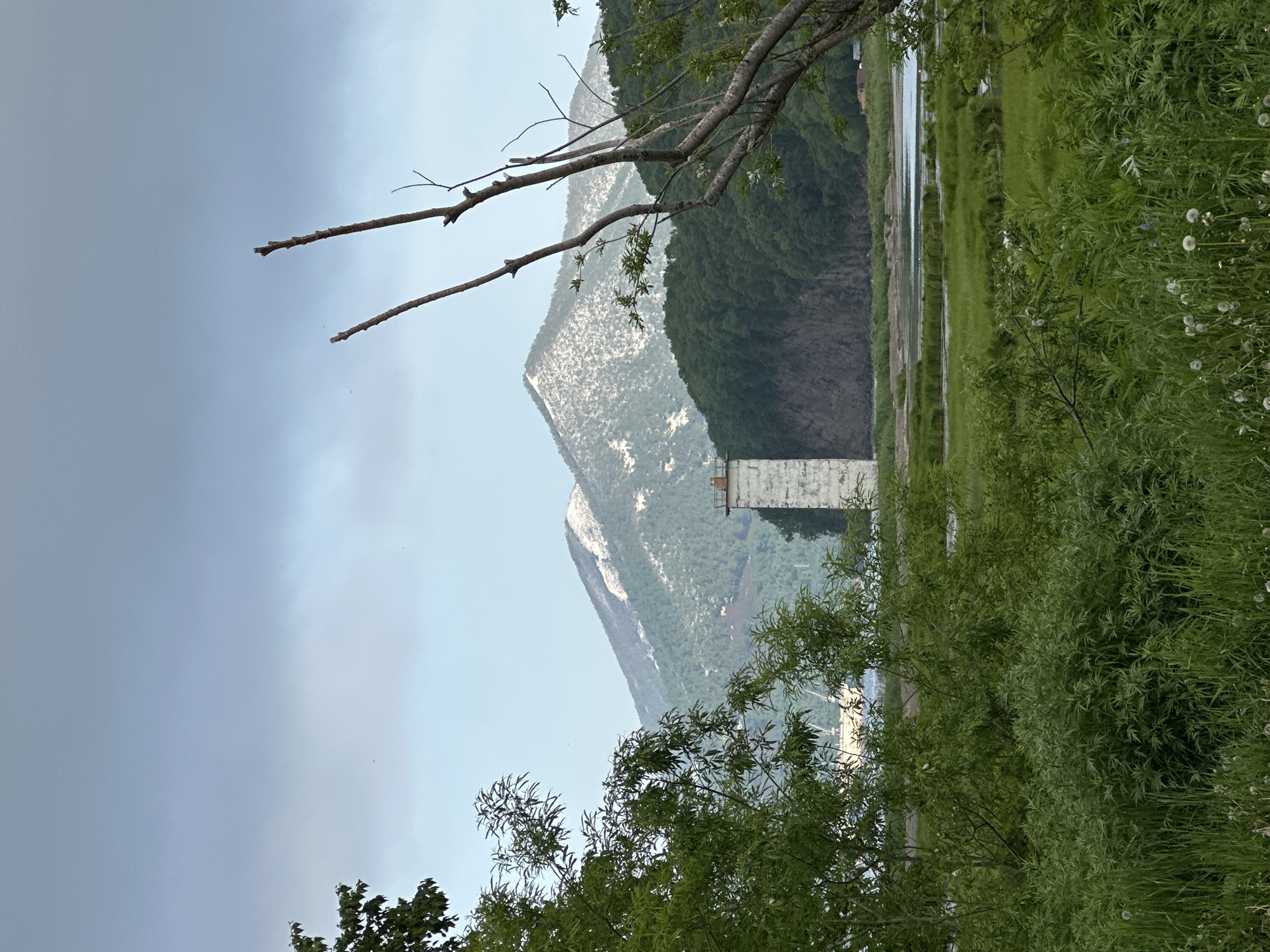 The image shows a serene landscape with a body of water in the foreground, likely the Avacha Bay, with lush green vegetation and trees on the banks. In the middle ground, a stark, white, rectangular structure, possibly a former industrial building or observation tower, stands near the water. Behind it, a steep, tree-covered hill rises, and beyond that, a large, snow-dusted mountain dominates the background, characteristic of the volcanic landscape around Vilyuchinsk, Russia, a city known for its proximity to active volcanoes. The sky is overcast with a soft, diffused light, suggesting a cloudy or misty day. The scene evokes a sense of natural beauty and a touch of industrial history.