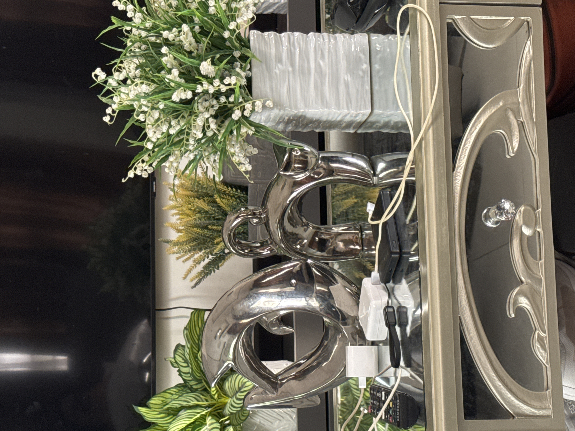 The scene depicts an interior setting, identified as being in Davao City, Philippines. A large, dark flat-screen television with a "SAMSUNG" logo on its lower left frame dominates the background, displaying reflections of an unseen light source or window and hints of distant foliage.
In the mid-ground, resting on a highly reflective dark surface, are several decorative and functional items. From left to right: a potted plant with broad, striped green and light green leaves, housed in a white textured pot; a polished silver metallic sculpture of two stylized dolphins or fish interlocked to form a dynamic, rounded shape; a smaller potted plant with fine, feathery yellow-green foliage; a smaller polished silver metallic sculpture forming an abstract arch or loop; and a bouquet of artificial white lily-of-the-valley flowers with green leaves, arranged in a white, horizontally ribbed ceramic vase.
The reflective dark surface, likely glass, mirrors the objects above it. Scattered across this surface in the foreground are various electronic accessories: a black Samsung remote control, a white rectangular USB wall adapter, a small white rectangular electronic case (possibly for earbuds), a black fitness tracker or smartwatch band, a black external hard drive or power bank, and multiple white and light-colored cables interconnecting these devices.
The objects rest on a console table, the front of which is visible in the lower portion of the image. The table features a light gold or silver-painted drawer front with a central arched cutout revealing a dark interior. Within this cutout, a stylized, wave-like metallic design is incorporated, and a clear, faceted crystal knob adorns the drawer. The very bottom edge of the image shows a partial view of what appears to be a person's legs or feet beneath the table.
