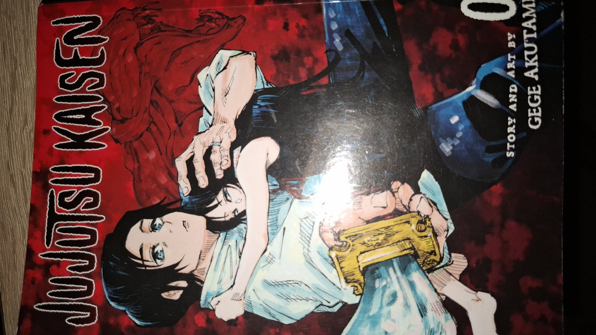 The image shows the cover of the manga "Jujutsu Kaisen 0" by Gege Akutami. The artwork features a character with black hair and blue eyes, wearing a light-colored robe and holding a weapon. A dark figure looms behind them. The background is a mix of red and dark blue hues. The text "Jujutsu Kaisen" is displayed vertically on the left.
The image does not contain information that can be used to identify the location being Sofia, Bulgaria.