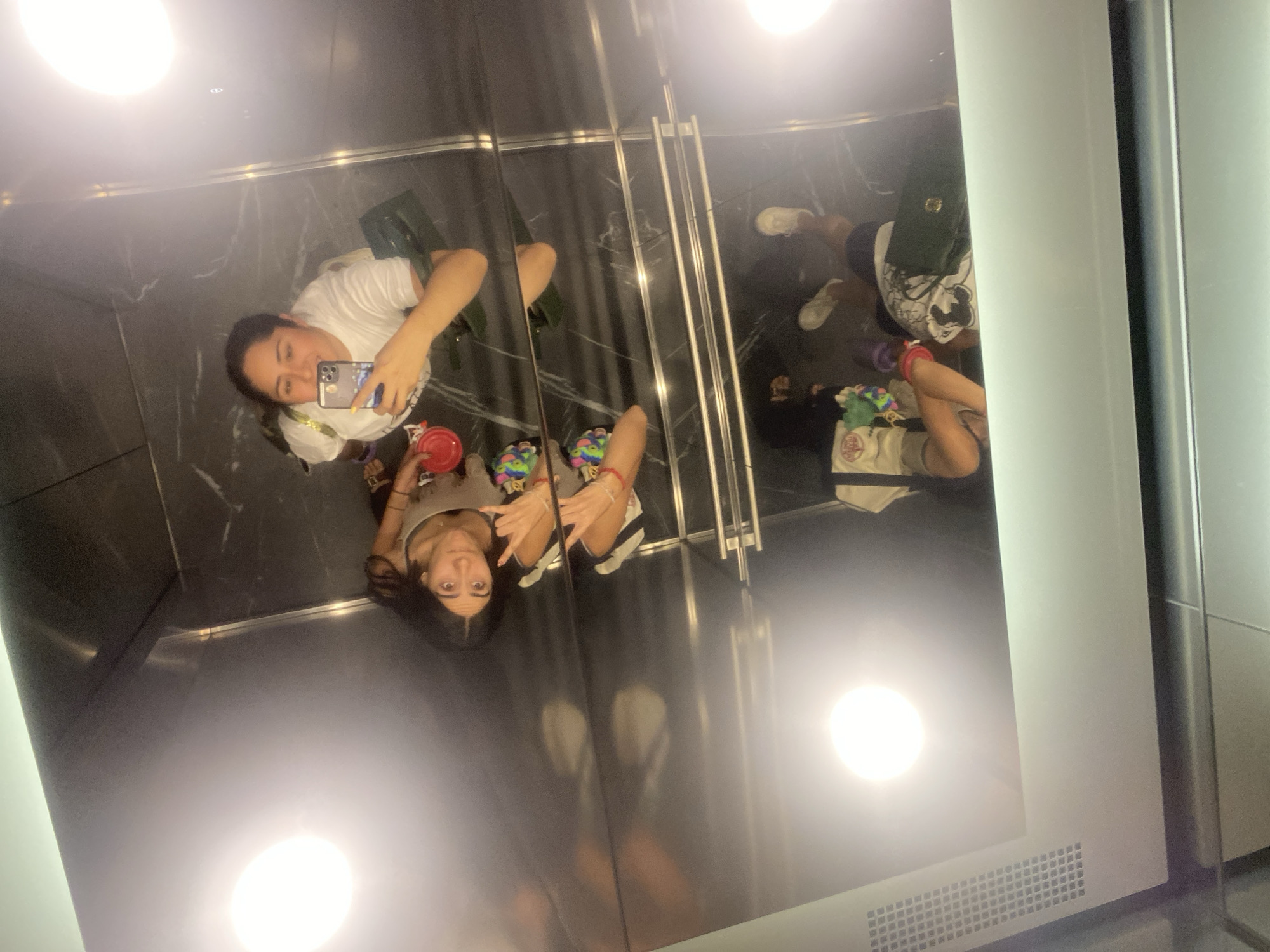 Two women are in an elevator with mirrored walls, with the reflection creating the illusion of multiple people. One woman wears a white t-shirt and is taking a picture with her phone. The other woman wears a grey tank top and is making a hand gesture. There is a red cup on the floor near the woman in the white t-shirt. Other objects on the floor include shoes, bags and other personal items. Based on the environment, this could be in a modern building in Puebla, Mexico.