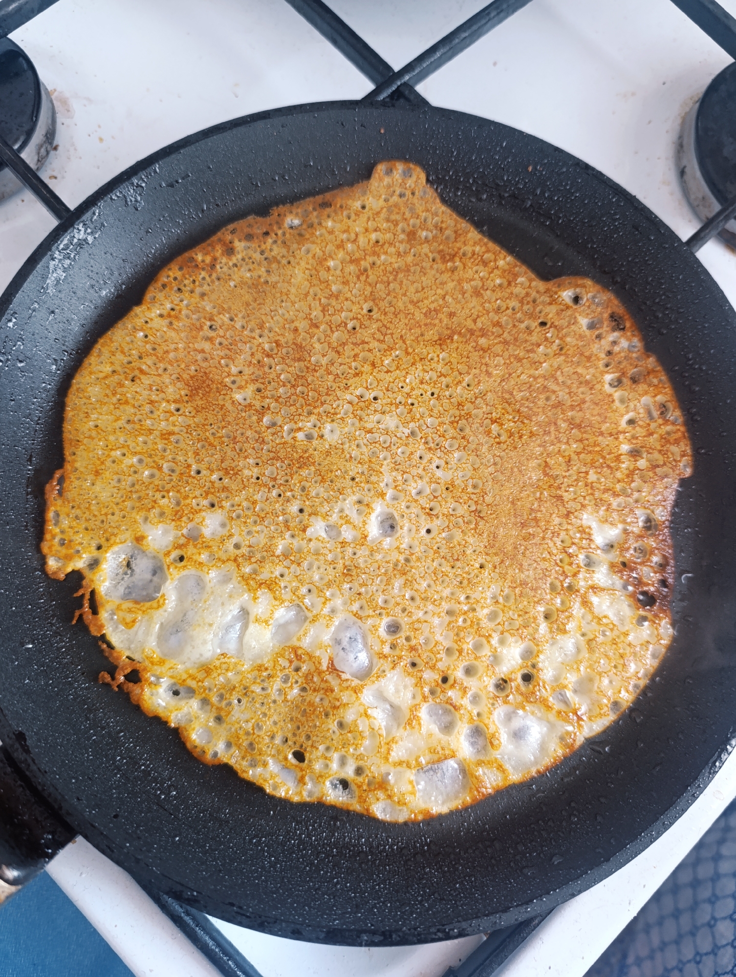The image shows a pancake being cooked in a black frying pan on a gas stovetop. The pancake is circular, with a golden-brown color and small holes. The lower part of the pancake appears less cooked and is white in color. The gas stovetop has a white base and black grates. The location is Unknown, Unknown.