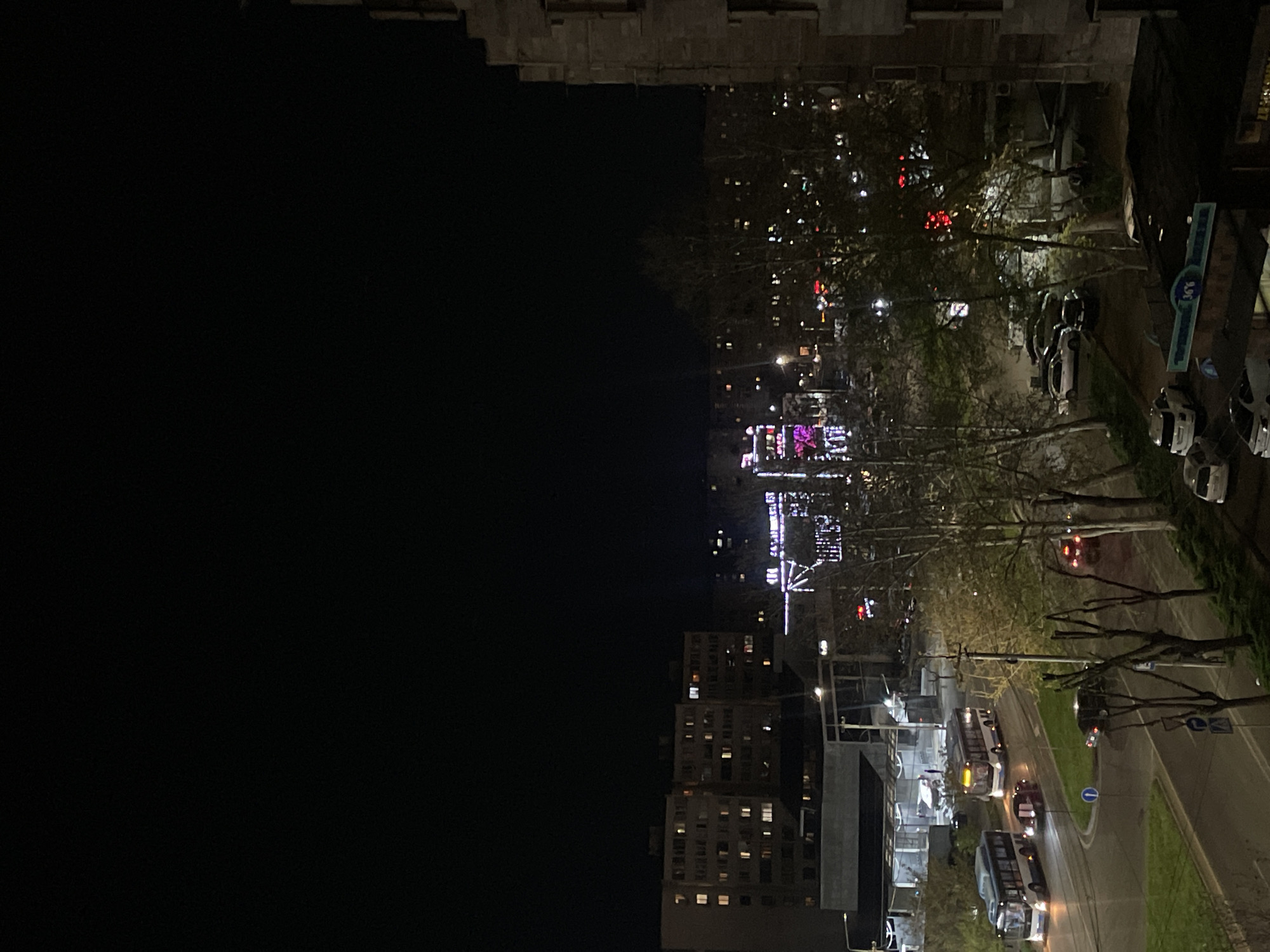 A night view from a high vantage point over Yerevan, Armenia. The lower portion of the image shows illuminated apartment buildings, streetlights, and traffic including buses and cars moving along a road. A park area with bare trees is present, along with a Ferris wheel illuminated with purple lights in the distance. Parked cars and buildings with illuminated signs are seen along the right edge of the frame. The upper-left portion of the image is dominated by darkness, likely a building close to the vantage point.