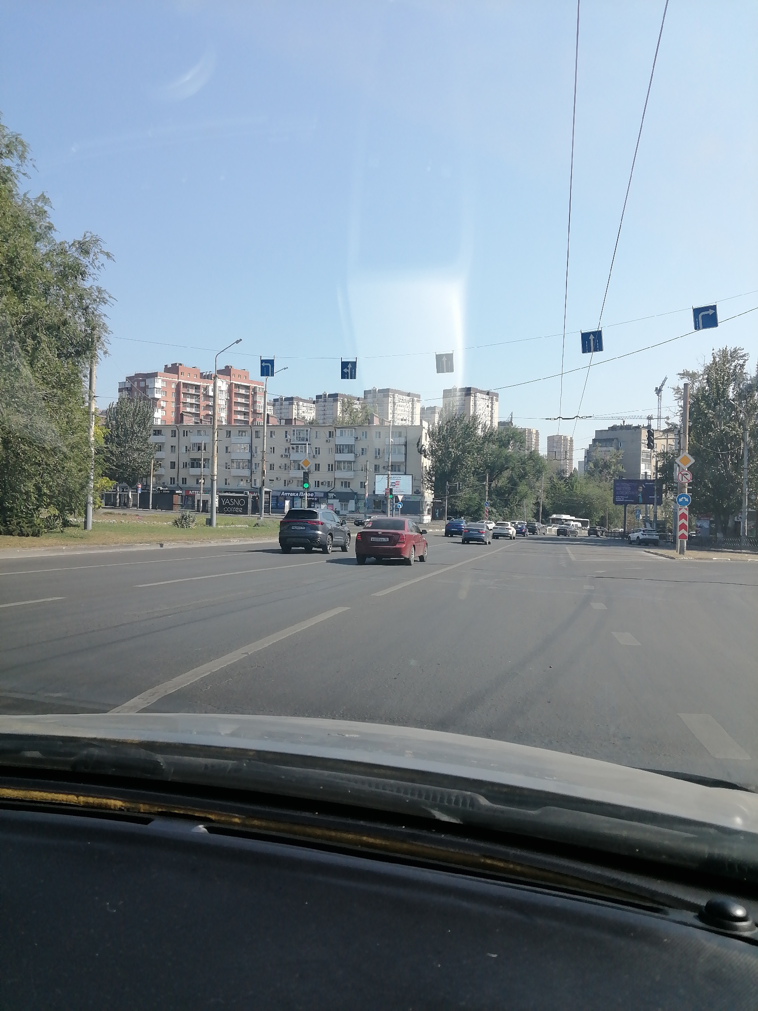 The image shows a view from a car's windshield looking forward at a multi-lane city street on a clear, sunny day. The road has lane markings and traffic signs overhead. Several cars are visible on the road, some moving and others stopped. In the background, there are tall apartment buildings, trees, and traffic lights. One traffic light displays a green signal. The perspective suggests the car is approaching or at an intersection. The urban environment appears to be a residential or mixed-use area within a city.