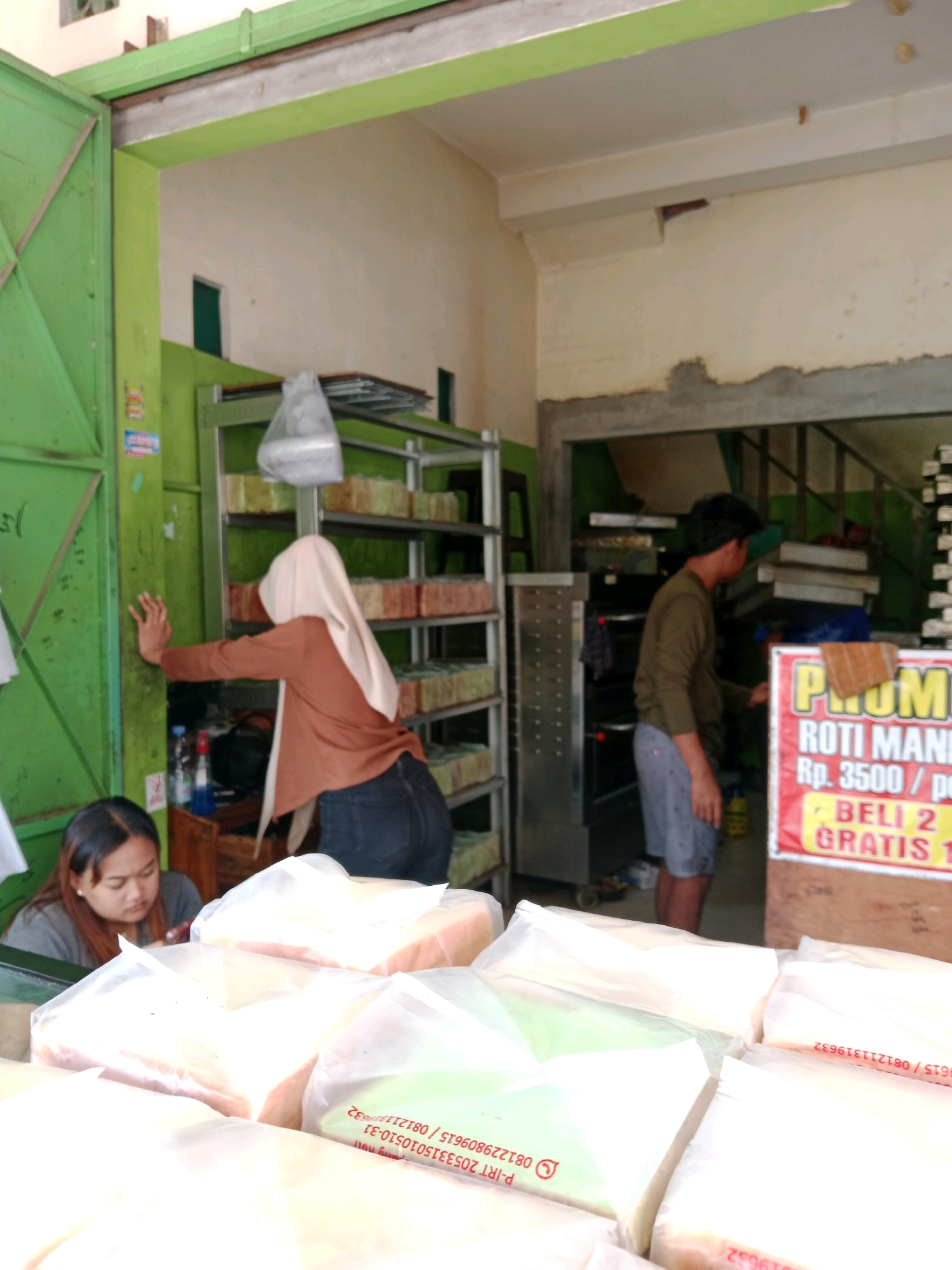 In Gentan, Indonesia, a bakery is depicted with a woman wearing a hijab and a brown top leaning against a green metal door. In front of her, another woman with dark hair is seated, looking down at something in her lap. Shelves stacked with loaves of bread wrapped in plastic are visible behind the woman in the hijab. To the right, a man in a green shirt and shorts is standing near an oven, and a red promotional sign displaying "PROMO ROTI MANIS Rp. 3500 / PC BELI 2 GRATIS 1" is prominently displayed. The environment suggests an active retail space with baked goods for sale.