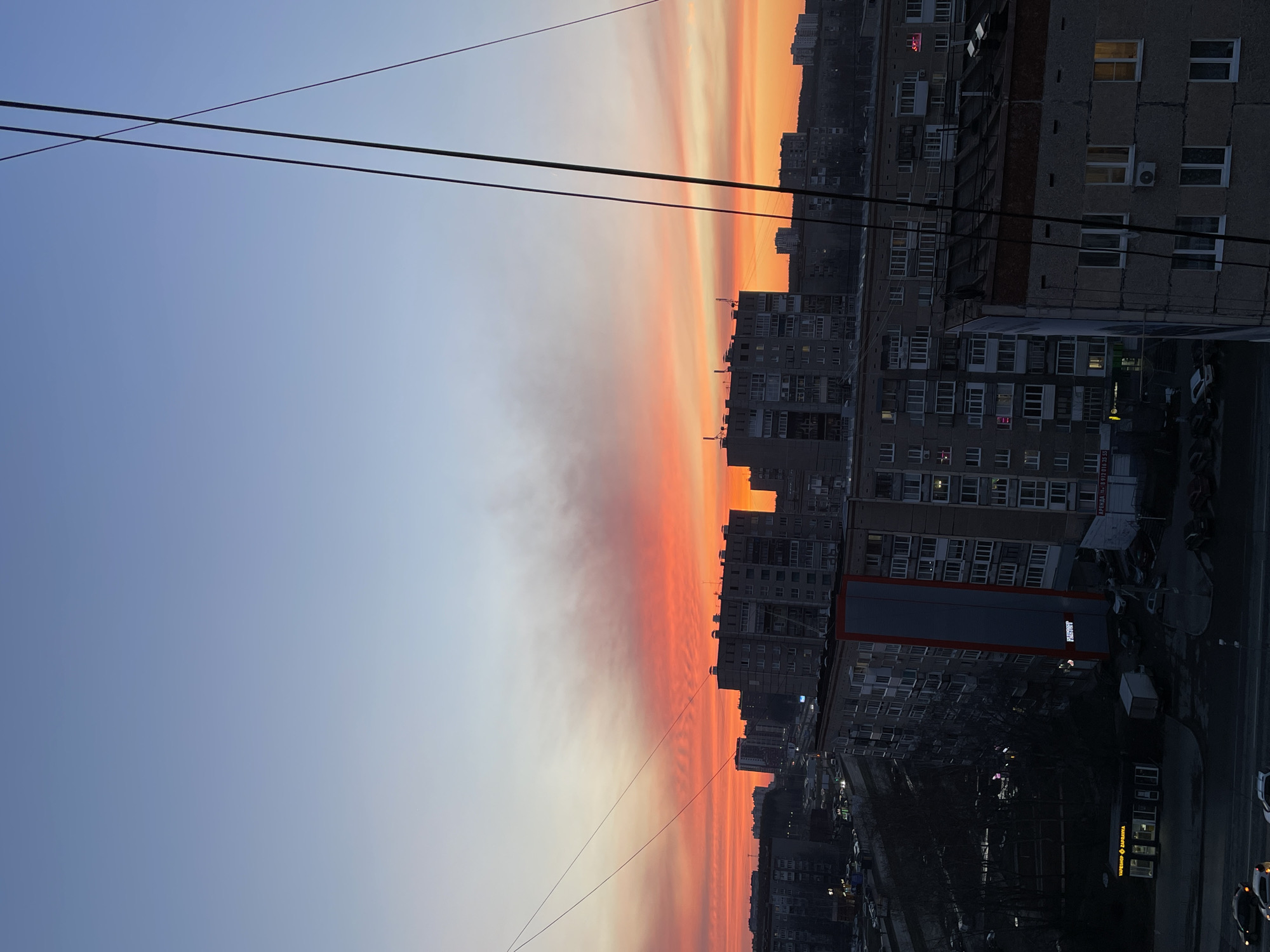 The image shows a view of a cityscape at sunset or sunrise. The sky is transitioning from a light blue to a vivid orange and red gradient. Several tall, gray apartment buildings are visible, densely packed together, suggesting an urban setting. Wires run across the upper part of the frame, intersecting the sky. The light on the buildings indicates that it is either early morning or late evening. The general architectural style of the buildings suggests a post-Soviet urban landscape. The scene likely comes from Izhevsk, Russia.
