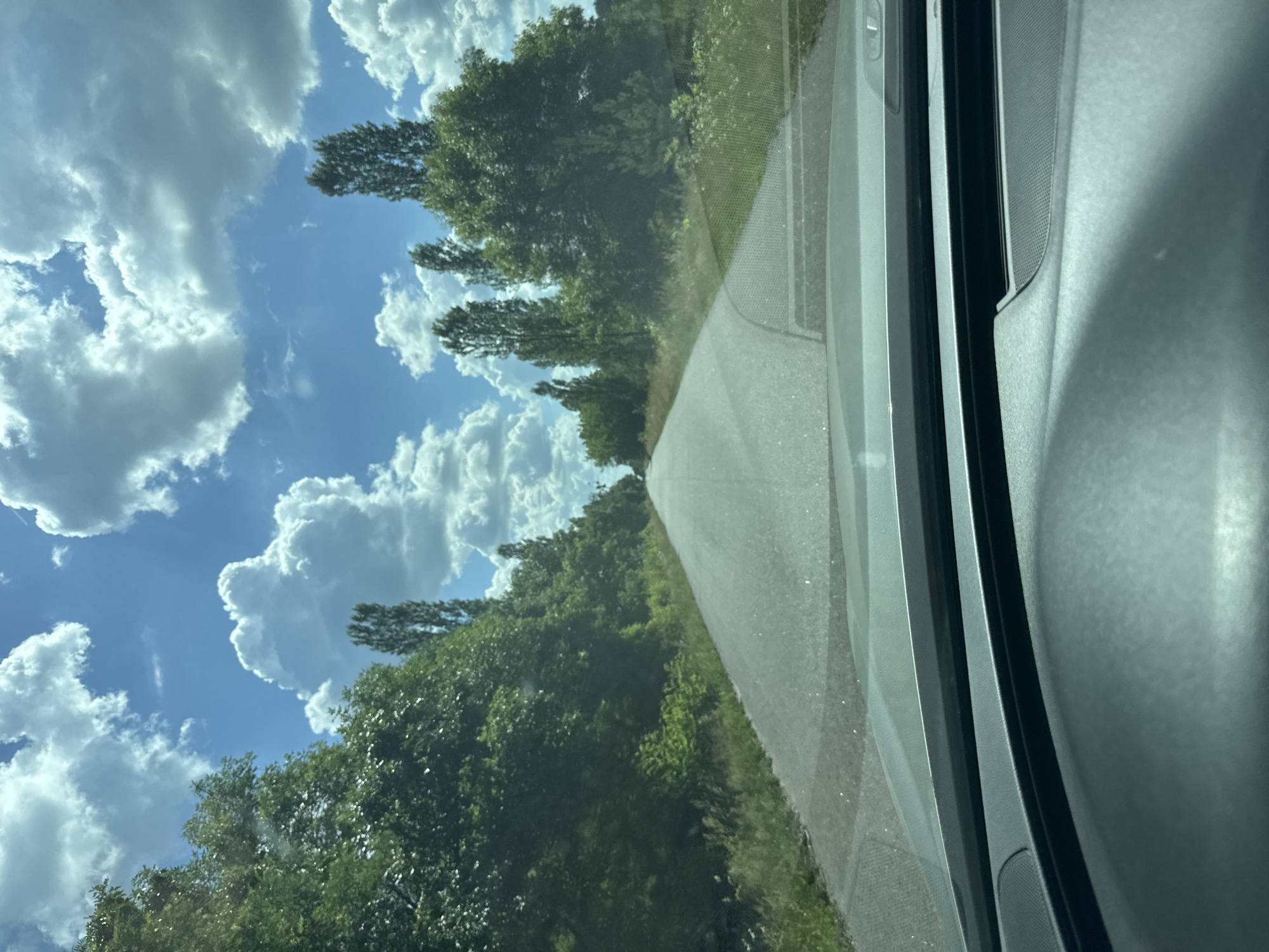 A first-person perspective shot from inside a vehicle shows a paved road disappearing into the distance, lined by dense green trees and foliage. The sky is a bright blue, dotted with large, fluffy white clouds. The dashboard of the vehicle is visible in the foreground. This scene is located on a road in the village of Dovhe, Ukraine.