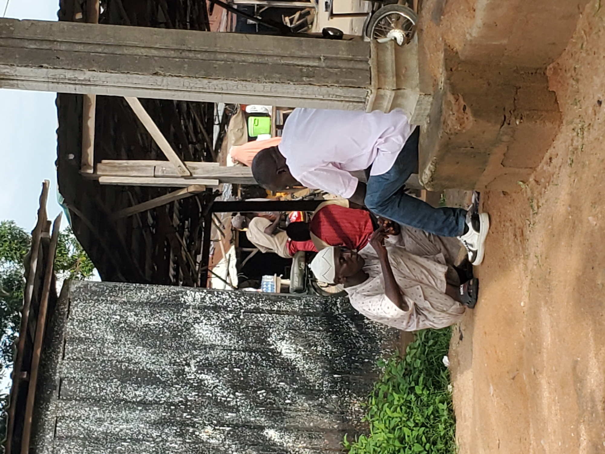 The image, taken in Mararaba, Nigeria, depicts an outdoor scene during the daytime, likely in a street or market area. The composition features several men engaged in a casual interaction.
In the foreground, a dark-skinned man wearing a light pink long-sleeved shirt and blue jeans is bending forward, looking downwards. He is wearing white and black sneakers. To his left and slightly in front, another dark-skinned man is seated directly on the reddish-brown dirt ground. He is dressed in a light-colored, possibly off-white, traditional long garment and a white prayer cap (kufi). He appears to be holding a small object, possibly a mobile phone, in his hands. The brand "NIKE" is faintly visible on the sole of his black sandal.
Behind the bending man, a person in a red shirt is partially visible, and further into the mid-ground, other figures and various goods are vaguely discernible within what appears to be a covered stall or market structure constructed with wooden beams and concrete elements. A prominent, dark, textured wall or surface occupies the left side of the frame. In the upper right corner, part of a bicycle wheel can be seen. Green foliage grows in patches on the ground, which is primarily bare earth. The bright sky in the background suggests clear weather conditions. The scene conveys a sense of everyday activity in a local community setting.
