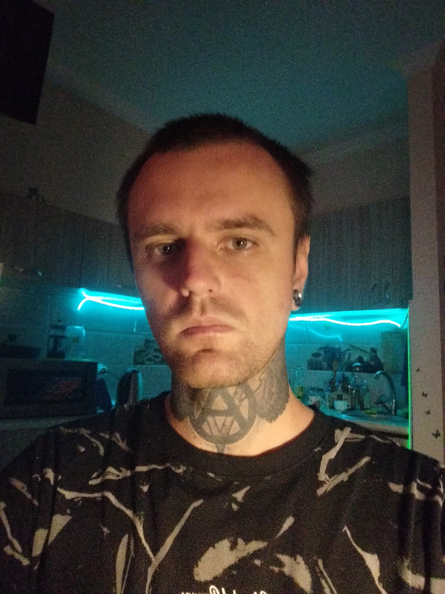 A close-up, selfie-style photograph shows a man with short brown hair, light skin, and stubble. He has a black tattoo of a pentagram on his neck. He is wearing a black t-shirt with a grey, abstract pattern. He has a grey ear expander in his left ear. The background appears to be an indoor setting, possibly a kitchen, with cabinets and a bright blue neon light strip running along the top of the cabinets. The lighting is dim, with the neon light being the primary source of illumination in the background, casting a blue glow. The overall impression is a dimly lit, casual indoor environment. This image does not contain any identifiable landmarks or features that would allow for specific location within Batumi, Georgia.