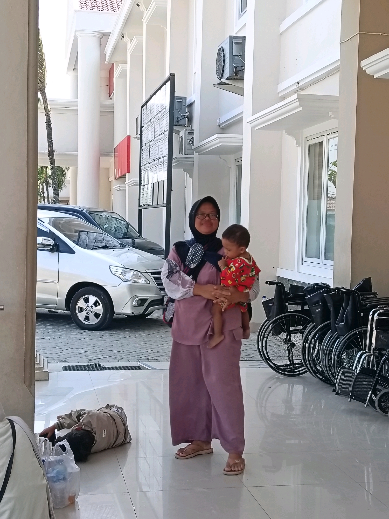 A woman wearing a pink hijab and glasses is holding a baby. The woman is wearing a pink outfit and sandals. The baby is wearing a red outfit with a pattern. They are standing in front of a building with white pillars and red accents. To the right of the woman are several wheelchairs lined up. In the background, there are cars parked. In the foreground, to the left, a person is lying on the floor next to some plastic bags. The scene appears to be in a hospital or medical facility. The city of Balun, Indonesia, is mentioned, but no specific landmarks or indicators of this location are visible in the image.