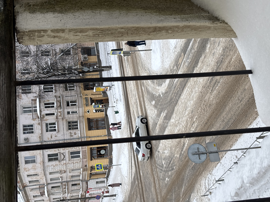 An elevated, downward-angled view captures a snow-covered intersection in Velikiye Luki, Russia, likely the junction of Prospekt Lenina and Oktyabr'skaya Ulitsa, centered on the building at 19 Prospekt Lenina. The foreground is partially obscured by three black vertical metal railings and a concrete pillar on the right, with accumulated snow on the immediate lower edge.
The scene depicts a winter day, with snow covering all sidewalks, verges, and rooftops. The road surfaces at the intersection are a mix of dirty slush, exposed pavement, and visible tire tracks, indicating vehicular traffic. A multi-story, light-colored corner building with classical architectural elements and signs of exterior wear dominates the background. Its ground floor, painted ochre, hosts businesses, including one marked "ПИВНИЦА" (Brewery/Beer House).
Four individuals are present:
1. Two persons, one in a pink coat and one in a darker coat, stand together on the snow-covered sidewalk near the "ПИВНИЦА" entrance, appearing to interact.
2. A solitary person in a long, light-brown coat stands on the far left sidewalk, near a pedestrian crossing sign.
3. A male individual in a dark jacket and blue jeans stands on the right sidewalk, positioned near a pole with traffic signals.
A white sedan, consistent with a Lada Granta, is mid-traverse across the intersection, moving from right to left. Multiple street signs are visible, including several yellow diamond-shaped pedestrian crossing markers, a red "No Entry" sign (partially visible), and a circular sign with a cross-like symbol in the lower-center foreground. A green trash receptacle is positioned near the two individuals at the corner, and a yellow advertising stand is also present there. Bare deciduous trees are visible behind the buildings.