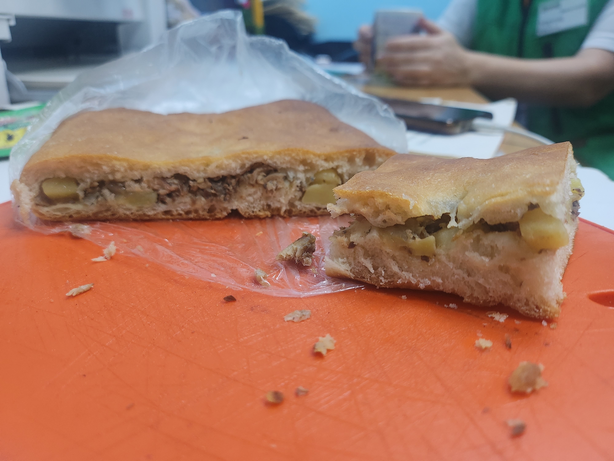 The image captures a casual indoor scene, primarily focusing on a large, savory pastry. The pastry, possibly a pie or pirog, is golden-brown and appears to have a thick crust. It has been cut into two pieces; a larger portion rests mostly within a clear plastic bag or wrap, while a smaller, triangular slice is separated and placed to its right on a bright orange cutting board or placemat.
The filling of the pastry is clearly visible in the cut sections. It consists of substantial chunks of light yellow potato and a darker, textured component that appears to be minced or shredded meat, possibly beef or pork. Some crumbs and small pieces of the pastry and filling are scattered on the orange surface around the food.
In the blurred background, a person is visible, wearing a green vest or apron over a white shirt. Their hands are clasped around a light-colored cup, suggesting they are drinking. The setting appears to be an office or breakroom, with indistinct objects like a desk or counter and potentially office equipment visible further back. No specific time of day or weather conditions can be discerned. There is no visible text in the image. The scene suggests a relaxed meal or snack break in Krasnogorskoye, Russia.
