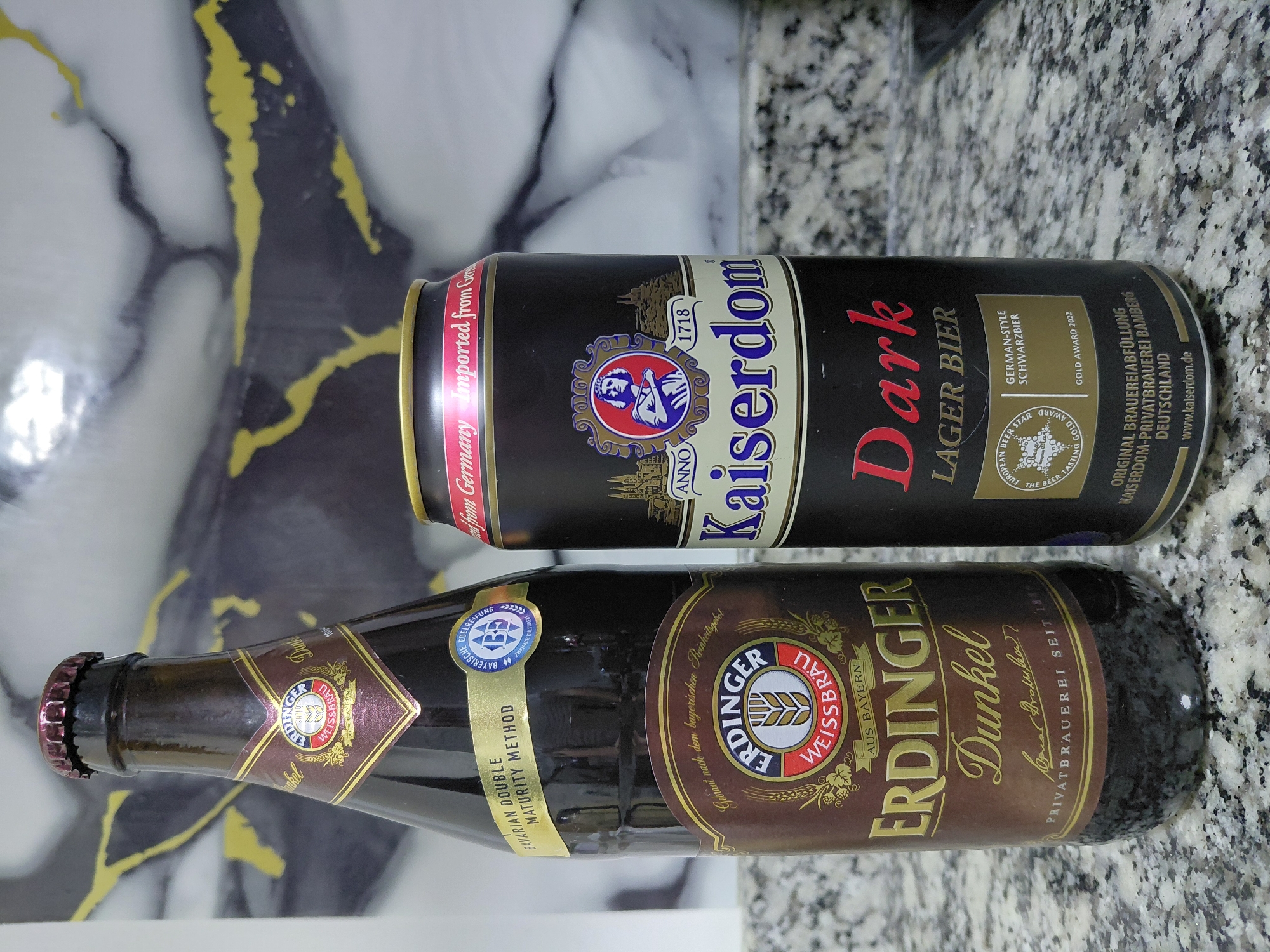 The image shows two dark beer containers, a bottle and a can, placed on a speckled granite countertop. The bottle, on the left, is a dark brown glass bottle with a metallic red cap. Its label is predominantly brown and gold, featuring the "Erdinger Weissbräu" logo with a wheat stalk and the text "Dunkel" in elegant script. Below this, it reads "Privatbrauerei seit 1886." The can, on the right, is black with a gold rim and red text. The prominent text on the can reads "Kaiserdom Dark LAGER BIER." There are also various award medals and text related to German origin and brewing. The background consists of a marbled pattern, likely a backsplash. The lighting suggests an indoor setting. The image does not contain any information that places it in Encarnacion, Paraguay.
