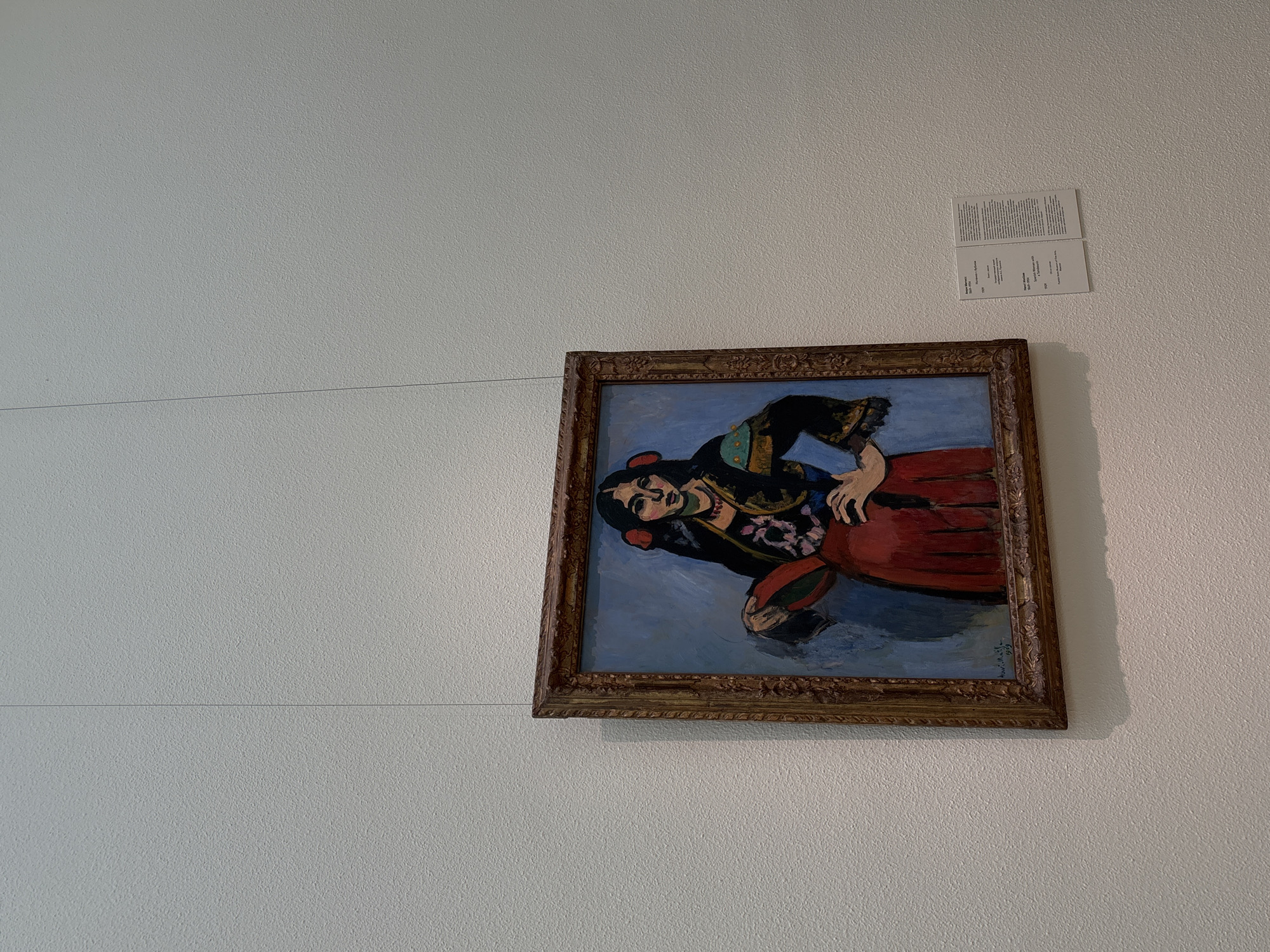 A color photograph taken at eye-level shows a painting hanging on a white wall with a small printed description of the artwork in Moscow, Russia. The painting, framed in an ornate gold-toned frame, depicts a woman in a red dress against a blue background. The woman is tilted sideways, and a thin cable runs horizontally across the wall in front of the painting. A small informational card is mounted on the wall above and to the right of the painting.