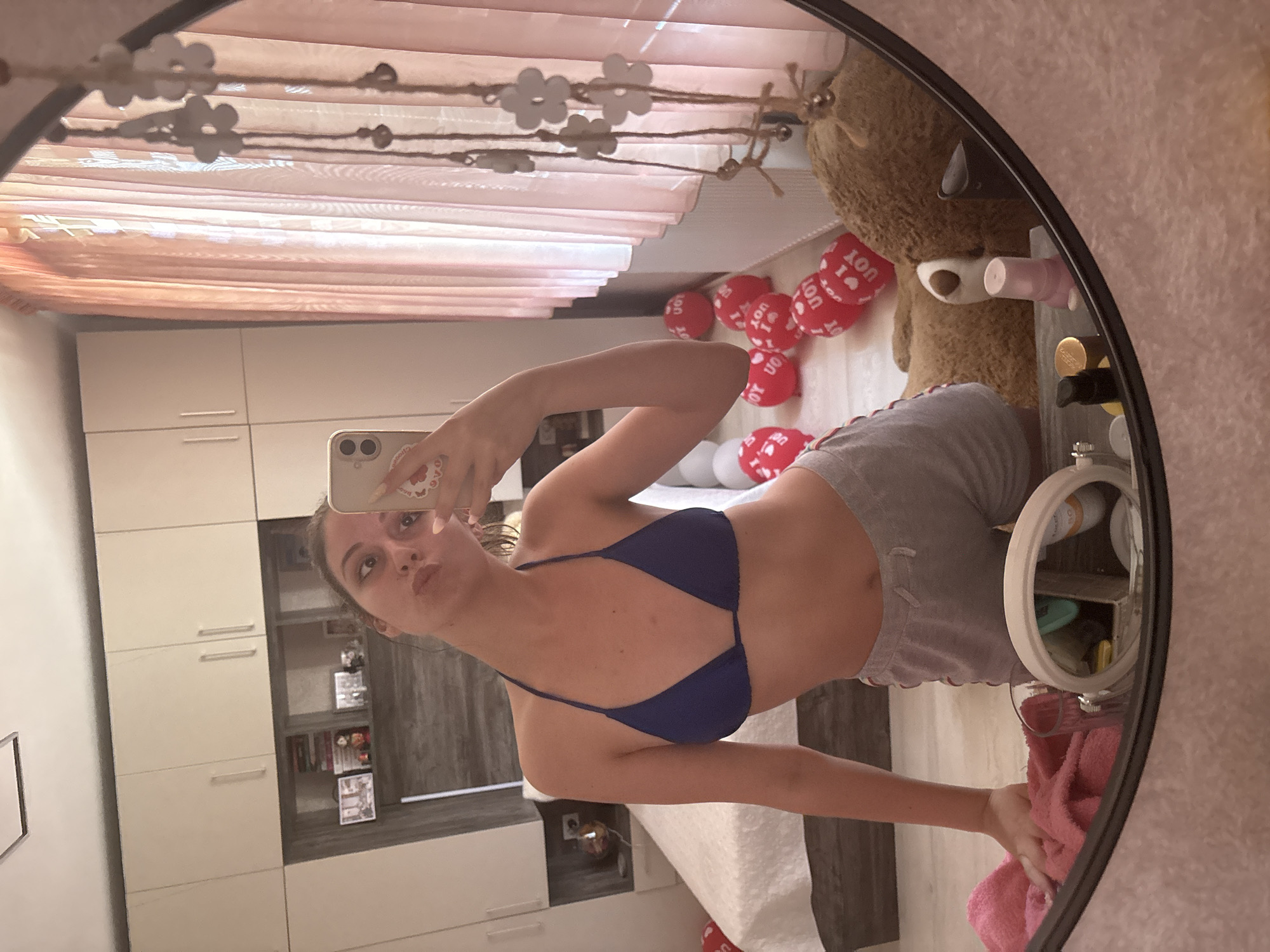 A young woman is taking a mirror selfie in a bedroom. She is wearing a blue bikini top and grey shorts. In the background, there are closets, a bed, and red heart-shaped balloons. There is also a teddy bear lying on the floor next to a dresser. The scene appears to be in a residential setting.