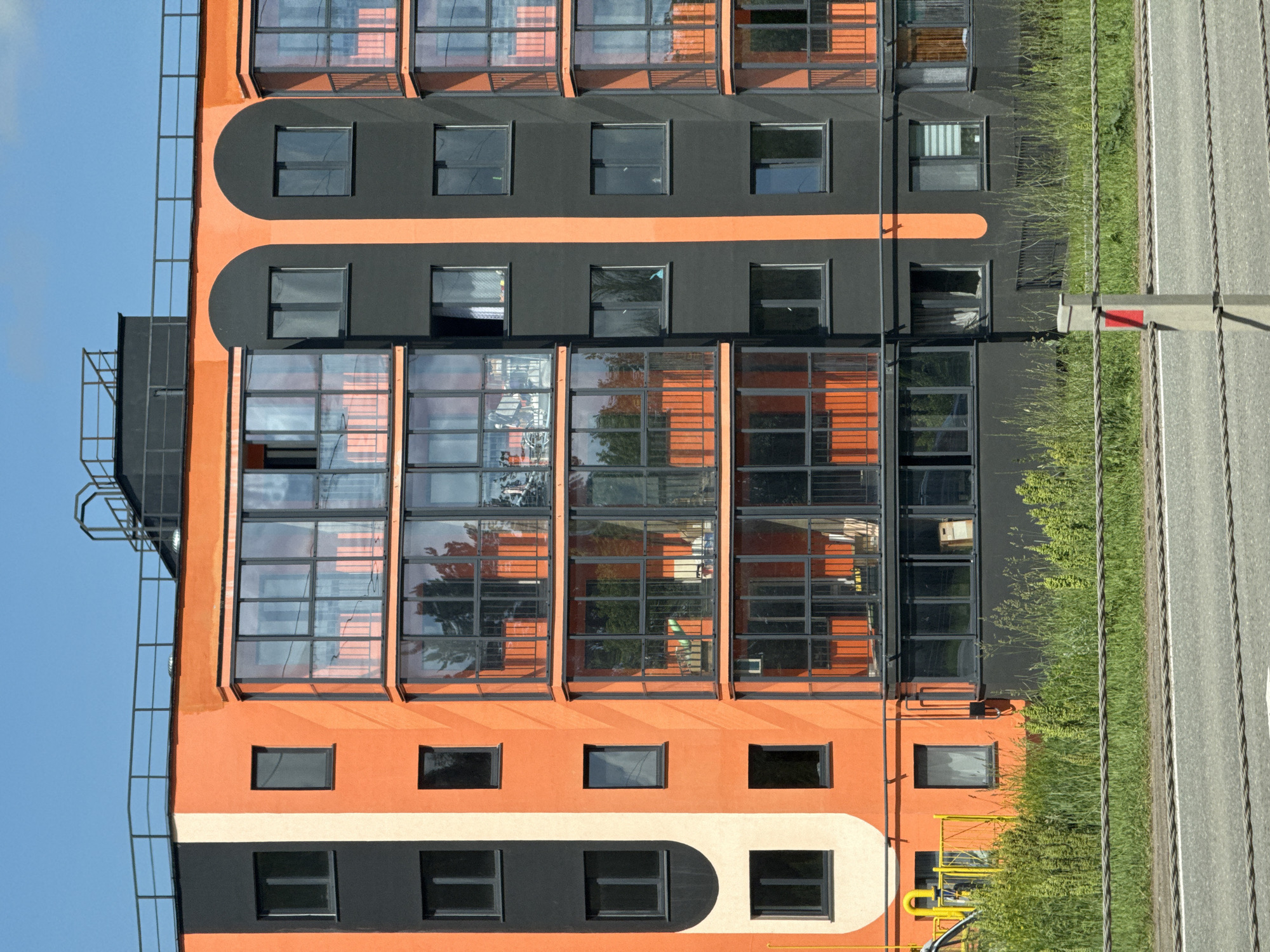 The image shows a modern apartment building with an orange and black facade, located in the city of Oktyabrsky, Russia. The building features multiple stories with large glass-enclosed balconies and dark-framed windows. The lower part of the building is surrounded by tall green grass and a road runs in the foreground, with steel cables stretched across it. The sky is clear and blue. There are no people visible in the image.