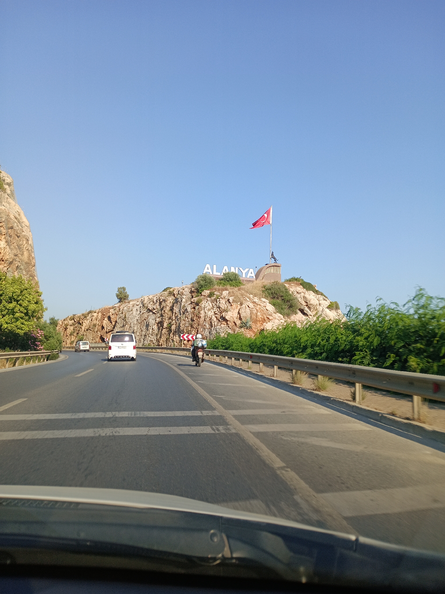 The image shows a view from a vehicle traveling on a road in Alanya, Turkey. On the right side of the road, there is a rocky hill with a large sign that spells out "ALANYA" in white letters. A Turkish flag is flying from a pole on top of the hill, behind the sign. The road itself is asphalt with lane markings, and there are other vehicles on the road, including a white van and a motorcycle. The surrounding environment appears to be dry and sunny, with sparse vegetation on the hillside and bushes along the side of the road. The sky is clear and blue.