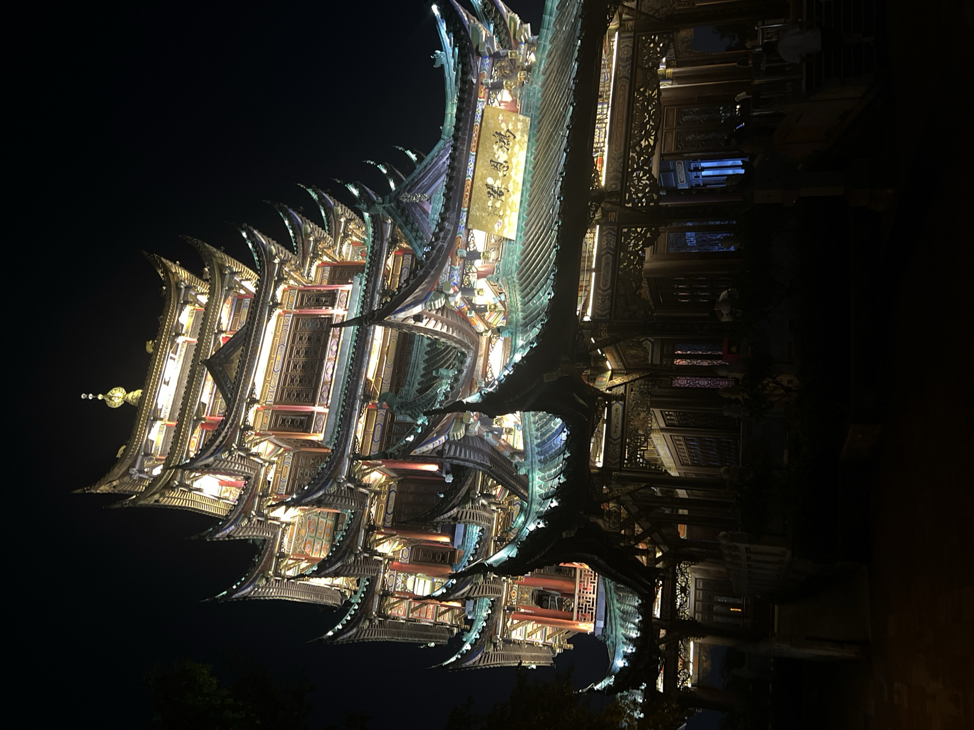 An illuminated multi-tiered pagoda-style building with intricate traditional Chinese architecture stands against a dark night sky. The building is lit from within, highlighting its ornate roofs with upturned eaves, colorful decorative elements, and vertical pillars. A large golden plaque with Chinese calligraphy is prominently displayed on one of the roofs. The foreground is dark, showing a pathway and some shadowed figures of people walking or standing. The scene appears to be an outdoor public space, possibly a temple or cultural landmark, at night. The location is identified as the Liangjiang district in China.