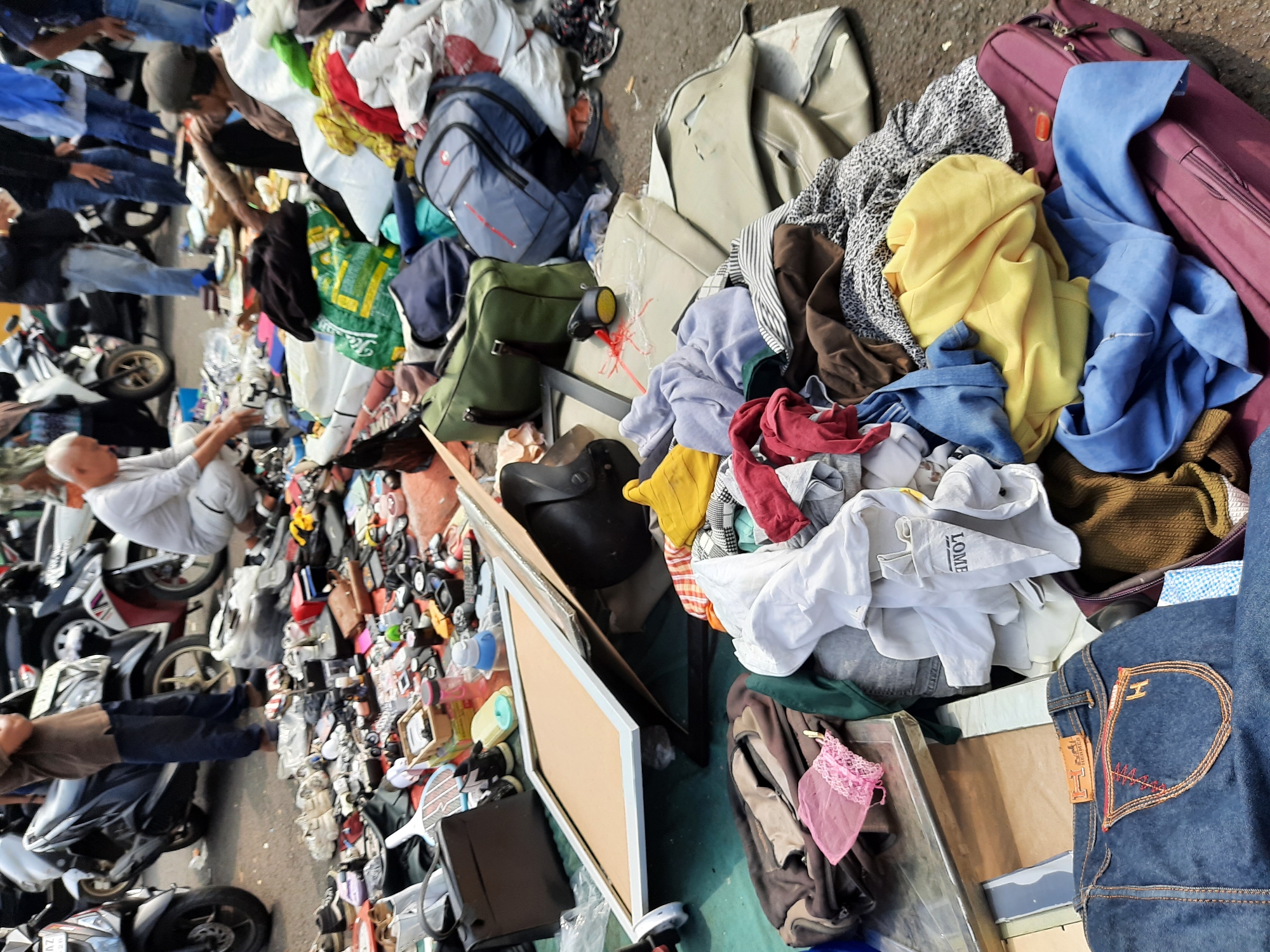 The image depicts an outdoor street market scene, likely in Bandung City, Indonesia, with a focus on a variety of second-hand goods. In the foreground, a pile of clothing is spread out on the ground, including jeans, t-shirts, and other garments. Some of the clothing items have visible brand tags or logos. Adjacent to the clothing are several bags, including a dark-colored handbag, a green duffel bag, and a blue backpack. A large framed object, possibly a picture frame, leans against a pile of items. Further back, more merchandise is visible, including shoes, boxes, and other miscellaneous items. Several motorcycles are parked in the background, suggesting the market is located on a street or open area. People are present in the scene, interacting with the goods, but their faces are not clearly visible. The ground appears to be asphalt or a similar paved surface. The overall impression is a bustling market with a wide array of affordable items for sale.