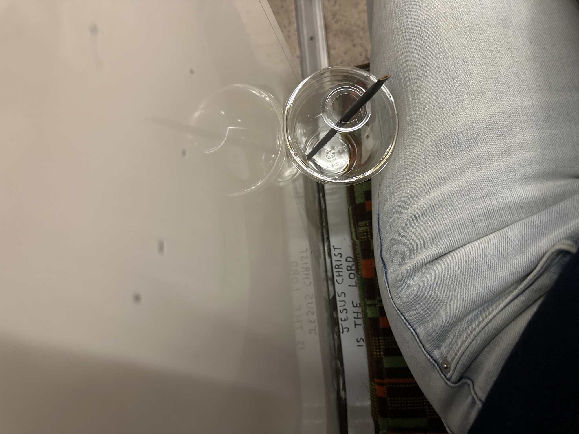 The image, taken in London, United Kingdom, presents an overhead view of an indoor scene featuring a clear plastic cup, a person's leg, and a reflective white surface.
In the mid-right of the frame, a clear plastic cup with a black straw rests on a surface. The cup appears to be nearly empty, with some residue at the bottom. Faint markings like "PP 5" and "LDPE," along with recycling symbols, are visible on its base. To the right of the cup, part of a person's leg, clad in light-wash blue denim jeans, is visible. The texture of the denim, seams, and a metal rivet are discernible. Below the jeans and to the left of the cup, a dark fabric with a multi-colored plaid or geometric pattern is present, possibly a bag, seat cushion, or carpet.
The left side of the image is dominated by a smooth, glossy white surface, likely a table or counter. On this surface, a white strip of paper or card displays clear, handwritten text: "JESUS CHRIST IS THE LORD". A prominent, reversed reflection of this text, appearing as "DROR SHT SI T2IRHC SUSEJ", is visible on the shiny white surface. The lighting is uniform, suggesting an indoor environment during the day. No specific activity or event is explicitly shown, beyond the arrangement of these objects.