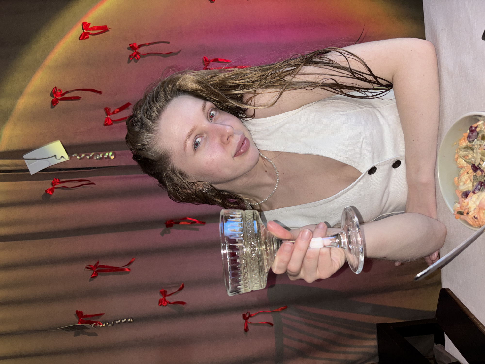 A young woman with long, wavy brown hair, wearing a light-colored sleeveless top and a pearl necklace, lies on her side, looking directly at the camera with a slight smile. Her left arm is bent, supporting her head, while her right hand holds a clear, ornate glass, possibly empty or containing a clear liquid.
The setting appears to be indoors, with a textured background that displays a gradient of warm colors, including browns, yellows, pinks, and reds. Scattered across this background are numerous small, red decorative bows, suggesting a festive or celebratory atmosphere. To the left of the woman's head, a small, white rectangular object, possibly an envelope or card with a black wavy line design, is visible next to a string of small, shiny metallic elements.
On the right side of the frame, a light-colored bowl contains a salad, which appears to be coleslaw or a similar mix with visible carrots, cabbage, and red fruit like cranberries or grapes. A metal utensil, likely a fork or spoon, rests beside the bowl on a light-colored tabletop or surface. The woman's relaxed posture and direct gaze convey a calm and possibly happy mood. There is no visible text in the image. The location context provided is Perm, Russia.