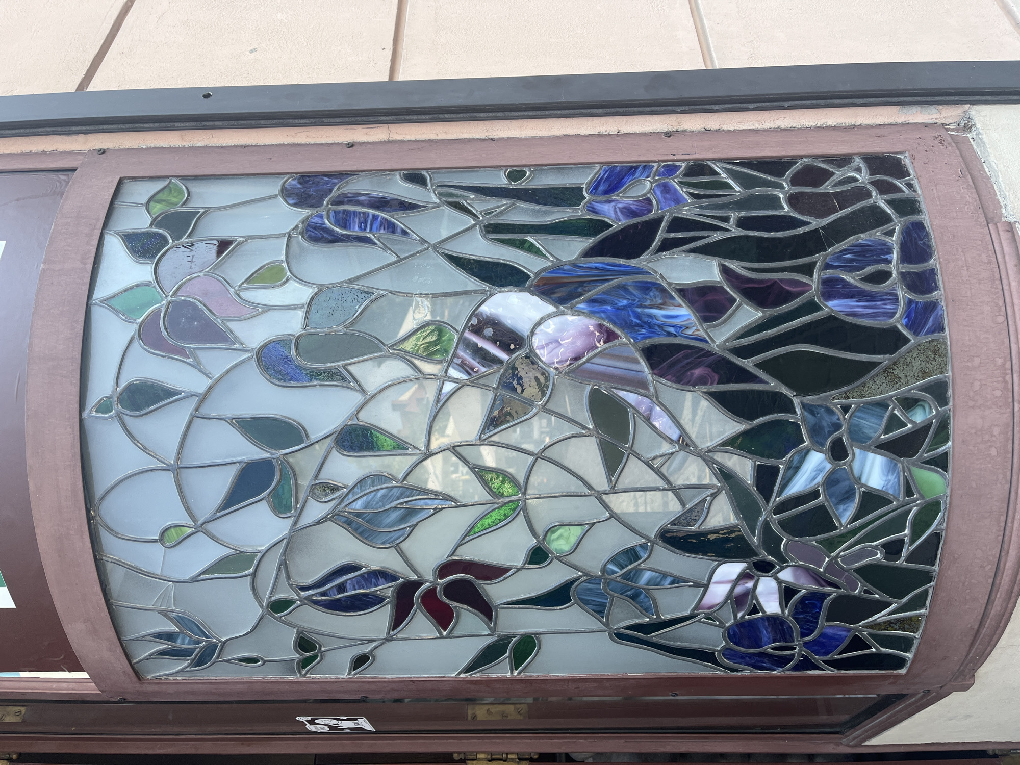 This media file shows a close-up view of a stained glass window. The window is framed with a dark brown material and is set against a textured, light-colored wall, possibly stucco or concrete. The stained glass design features an abstract floral or leaf motif, rendered in various shades of blue, purple, green, and grey, interspersed with clear and frosted glass. The pieces of glass are of irregular shapes and are separated by lead lines, typical of stained glass artistry. Some of the glass pieces have a swirling, marbled pattern, particularly the blues and purples. The overall impression is one of organic shapes and a somewhat muted color palette, with the clear glass allowing some light to pass through. The reflection of what appears to be the surrounding environment, including possibly an overhang or ceiling, can be seen on the surface of the glass. The window is installed in what seems to be an exterior setting, possibly a storefront or building facade, as suggested by the visible wall and the wooden frame. There is no discernible activity or event taking place; the focus is solely on the static artwork of the stained glass window. The time of day and weather are not clearly indicated, though the lighting suggests it is daytime. No visible text is present within the stained glass itself, though a faint white sticker or label is visible on the dark frame below the window.