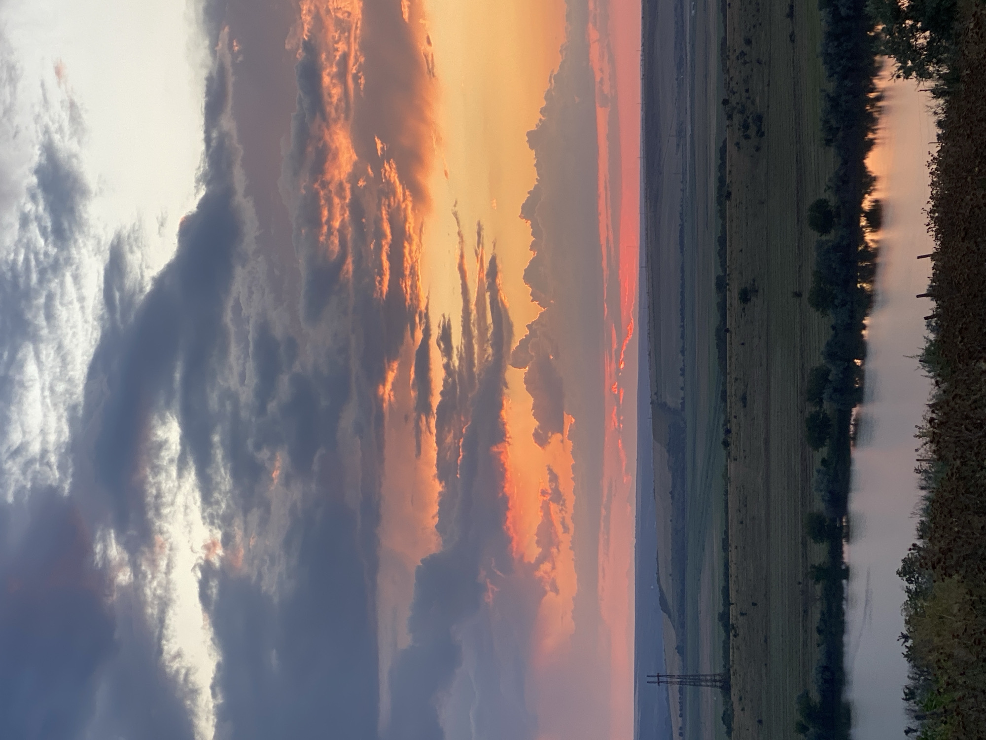 This is a photograph of a sunset over a rural landscape in Cairaclia, Moldova. The sky is filled with dramatic clouds painted in hues of orange, pink, and gray. A body of water, likely a river or lake, occupies the foreground, reflecting the colors of the sunset. Beyond the water, rolling hills and fields stretch towards the horizon, dotted with trees and agricultural land. A tall electrical pylon stands prominently on one of the hills. The overall scene evokes a sense of natural beauty and tranquility. There are no people or animals visible in the image, and no actions or interactions are taking place. The environment is a natural landscape characterized by its topography, vegetation, and the atmospheric conditions of sunset.