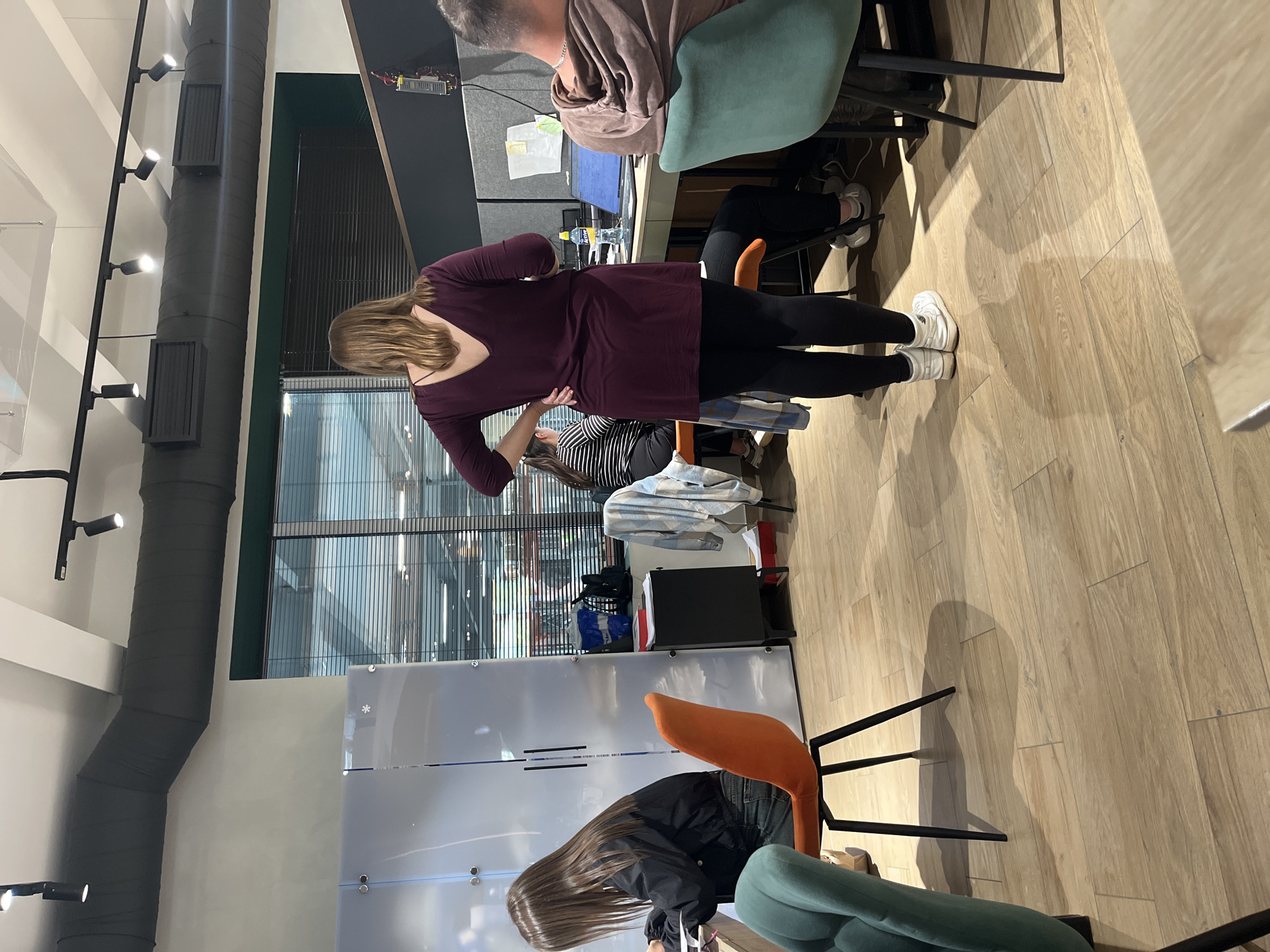 This image captures an indoor scene, likely an office or co-working space, populated by several individuals engaged in various activities. The perspective is from eye-level, looking across the room.
In the foreground, a woman stands with her back to the camera, wearing a dark purple long-sleeved top and black leggings paired with white sneakers. Her hands are positioned on her lower back, and her light brown hair falls over her shoulders. Directly behind her, another person is seated at a desk, their back visible, wearing a brown hooded sweatshirt. To the left, another individual with long dark hair is seen sitting, wearing a black jacket and jeans, looking downwards. A fourth person, partially obscured, is visible further back, wearing a striped top.
The setting features modern office furniture, including various chairs in colors like green and orange, and sleek black desks. A computer monitor is visible on one of the desks, along with a yellow-labeled water bottle that appears to have "ACTY" written on it. Papers and bags are scattered around the workspace. The floor is made of light-colored wooden planks, and prominent shadows from people and furniture are cast across it, suggesting bright overhead lighting or natural light from unseen windows.
Above, exposed dark grey air ducts run across the ceiling, fitted with several track lights illuminating the space. Large windows with vertical blinds or grates are visible in the background, suggesting an exterior view. A translucent partition or whiteboard with a small snowflake decal is also part of the room's decor. The overall atmosphere appears neutral and typical of a busy workspace during daytime hours.