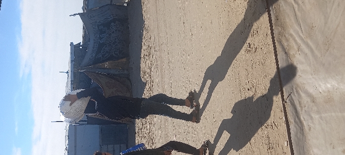 Two individuals walk left across a dirt and sand path in Khan Yunis, Palestinian Territory, under a clear blue sky. The central figure wears a dark long-sleeved top, dark blue jeans, and sandals, balancing a large, irregularly shaped white sack on their head, supported by their right hand. A second individual is partially visible on the left, also wearing dark clothing and sandals, with a blue item draped over their left shoulder. Long shadows from the individuals are cast on the light brown ground. In the background, several improvised structures made from corrugated metal, fabric, and tarps are visible, suggesting a makeshift settlement. A concrete or paved edge runs horizontally across the bottom foreground.
