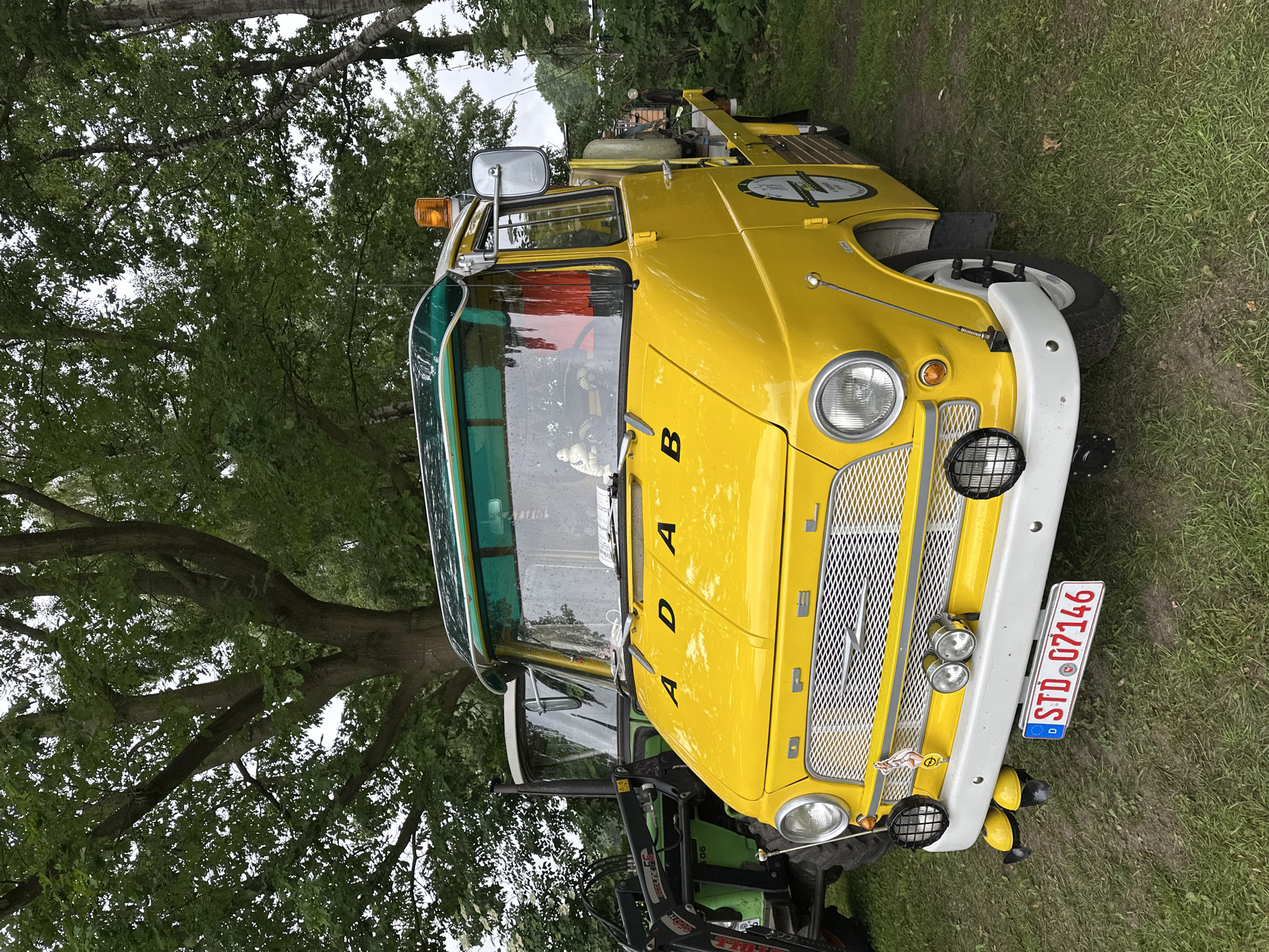 This image captures a well-maintained vintage bright yellow Opel Blitz utility truck, likely a service or tow truck, parked on a grassy field in Deinste, Germany. The vehicle is positioned in the foreground, facing slightly towards the left, showcasing its front and driver's side. The Opel Blitz features a cab-over-engine configuration with a glossy yellow paint finish and numerous chrome accents. The front grille is chrome with a prominent Opel "lightning bolt" emblem at its center and "OPEL" spelled out in chrome letters above it. Round headlights are present on both sides, with smaller, circular amber turn signals positioned below them. A white bumper extends across the front, supporting two mesh-covered black auxiliary lights on either side and two smaller, chrome auxiliary lights centrally mounted. A small pin-up girl decal is affixed to the right side of the bumper. Below the bumper, two yellow, rounded objects, possibly decorative covers or small horns, protrude downwards. The German license plate, displaying "D STD 07146" in red on a white background, is centrally mounted on the bumper. The letters "A D A B" are individually applied in black on the hood. An antenna extends from the front right fender. The windshield is large and slightly curved, with a green transparent sun visor extending across its top. Wipers are visible on the windshield, which appears wet, suggesting recent rain or condensation. The interior of the cab shows a red fabric element, possibly a curtain, and a small white figurine on the dashboard. A large chrome side mirror is mounted on the driver's side. An orange beacon light is affixed to the roof, above the driver's side. The truck's side panel features a circular black and white decal, partially obscured, with text that is not fully legible but resembles a service logo. The front wheel on the driver's side has a white steel rim and a black tire, indicating a robust design. The rear of the truck, partially visible on the right, shows a flatbed area equipped with a yellow cylindrical tank and other utility equipment. To the immediate left and slightly behind the yellow truck, a green and white agricultural tractor is partially visible. It features a prominent black front loader with "ECOLINE F" or similar text visible on its arm. The tractor's cab is white, and a dark exhaust pipe extends upwards. Part of a large front tire is visible. The ground consists