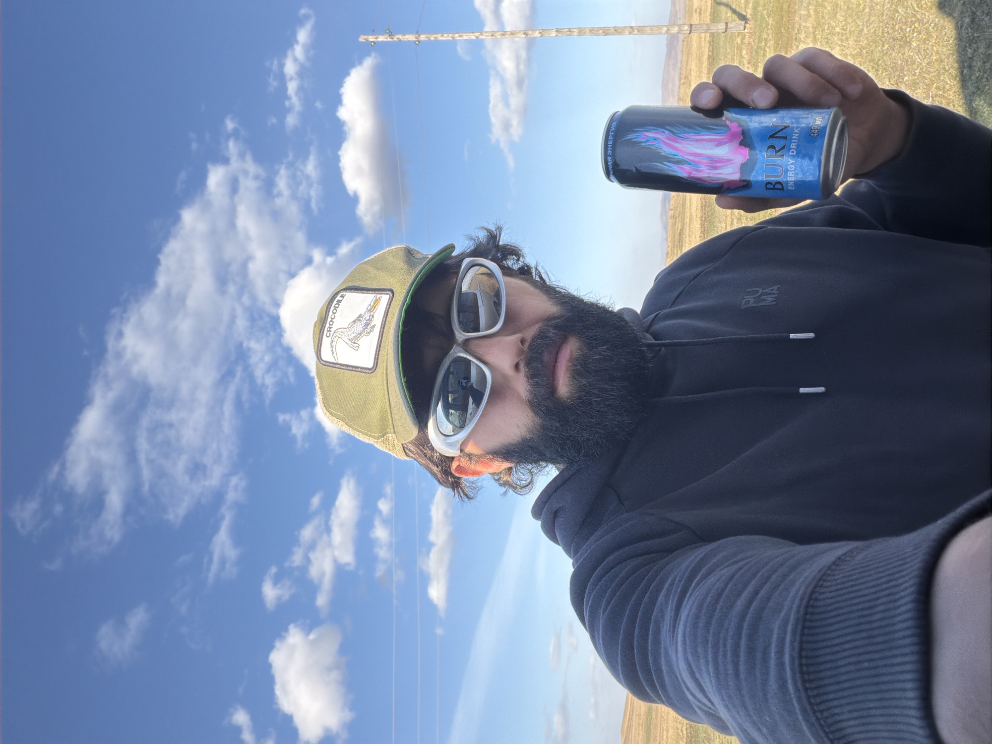 A selfie photo depicts a bearded man with sunglasses and a Crocodile-branded hat holding a "BURN" energy drink. The background reveals a landscape with a blue sky, white clouds, a grassy field, and a utility pole. It's daytime. There's no specific indication this scene is taking place in the city of Знаменка, Russia. The man wears a dark Puma hoodie.