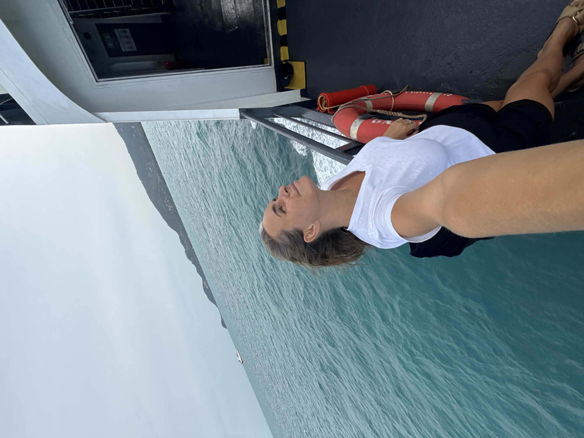 A woman with blonde hair is taking a selfie on the deck of a ferry. She is wearing a white t-shirt and black shorts. She is looking up at the sky with her eyes closed. The ferry is moving through turquoise water, and a distant island is visible on the horizon. The sky is overcast. The deck of the ferry is dark, and there is a red life preserver visible. The location is the Gulf of Thailand, near Ko Samui District, Thailand.