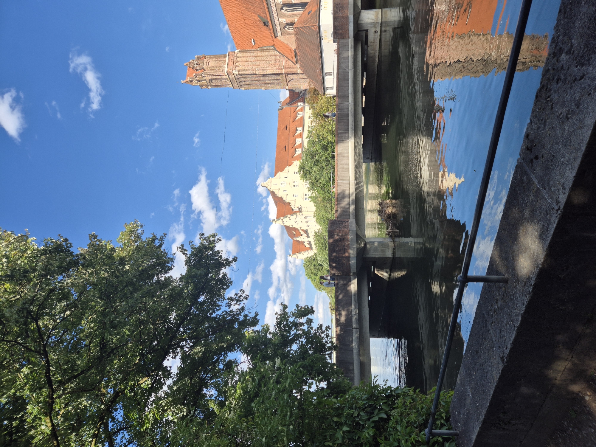 The image depicts a scene in Landshut, Germany, featuring a bridge crossing a body of water. On the right side of the frame, a tall brick tower with a red tiled roof rises prominently, adjacent to a building with a similar red tiled roof. To the left of the tower, a large, pale yellow building with multiple red gabled roofs and numerous windows is visible, nestled behind lush green trees. A concrete bridge spans the water, supported by pillars. The water reflects the sky, clouds, and the buildings and bridge above. In the foreground, a stone embankment with a black metal railing lines the water's edge. A large, leafy tree occupies the upper left portion of the image. Two individuals can be seen standing on the bridge, facing away from the camera, and another individual is walking on the bridge. The sky is bright blue with scattered white clouds.