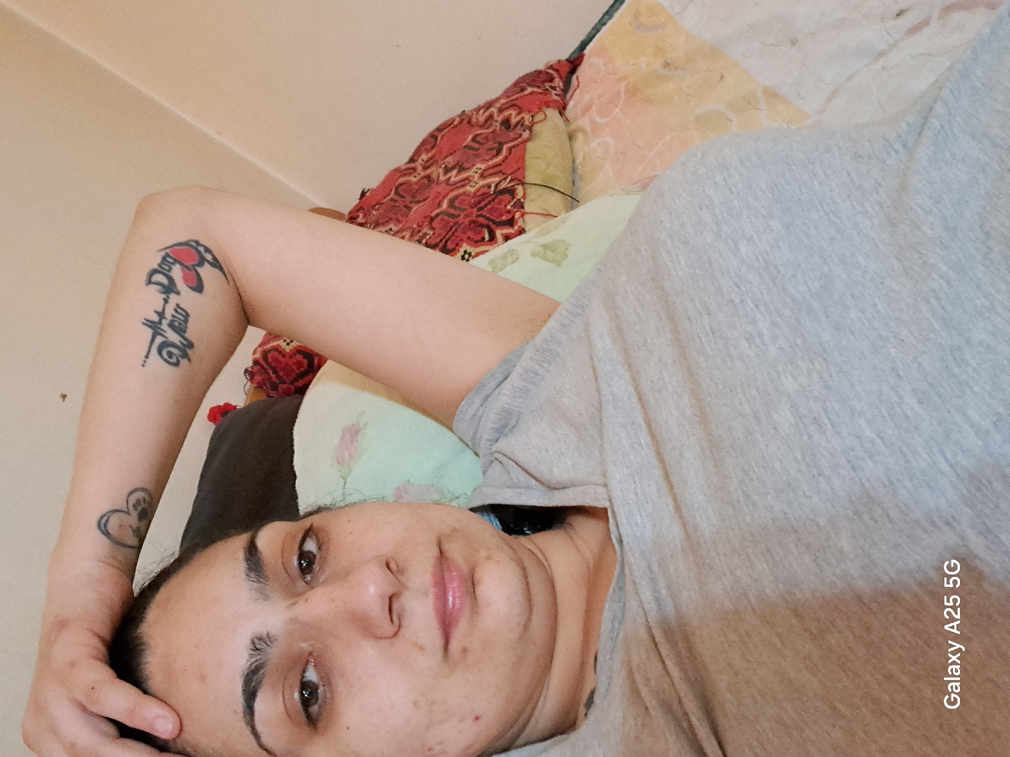 The image captures a person, likely a woman, lying on their side in an indoor setting, looking directly at the camera. She has medium-toned skin, dark hair, and prominent dark eyebrows. Her expression is neutral to contemplative, with her eyes open and her lips slightly parted.
She is wearing a plain light grey t-shirt. Visible on her left arm are two tattoos. On her upper arm, there is a striking tattoo of a red butterfly outlined in black, with intricate black script or a design beneath it. Further down on her wrist, a smaller tattoo depicts a heart containing a paw print.
The person is resting on a bed, indicated by several pillows with varying patterns and colors, including a vibrant red and black patterned one, a light green pillow with a faint floral design, and a light yellow or orange one. The background is a simple, light-colored wall, suggesting a bedroom or similar personal space. The overall lighting appears soft and natural, characteristic of daytime indoors.
In the bottom right corner of the original image, the text "Galaxy A25 5G" is visible vertically, identifying the device used to capture the photograph. Although the location context provided is Karnobat, Bulgaria, there are no specific visual cues within the image itself to confirm this geographical detail.